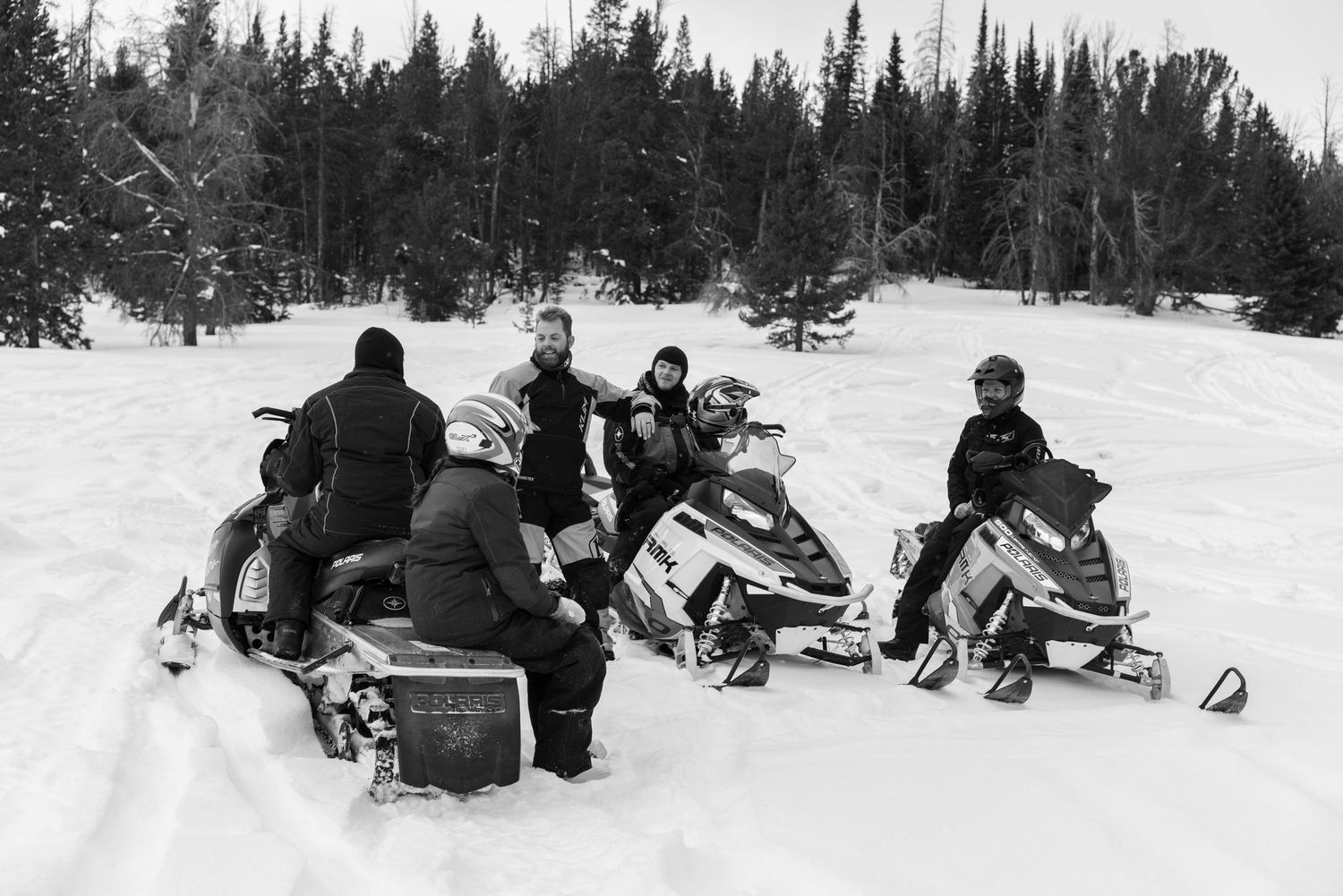 Group of snowmobilers resting in snowy winter landscape with forest backdrop, black and white photo.