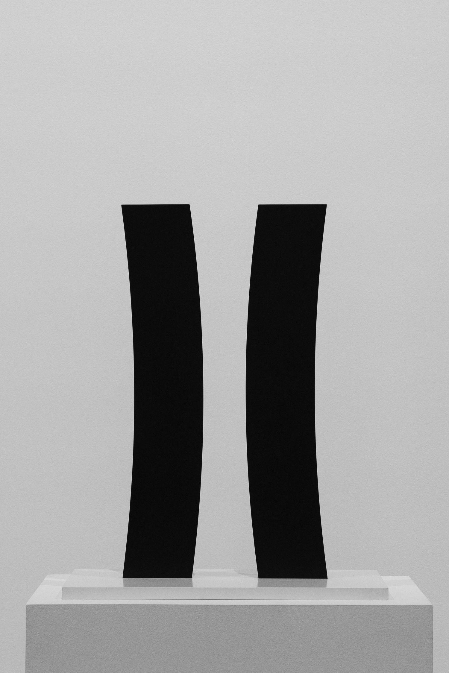 Two tall curved black vertical sculptures displayed on a white pedestal against a gray gallery wall.