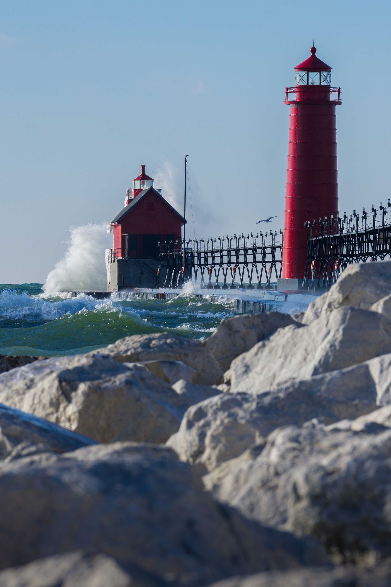 Bright red lighthouse and pier with crashing waves and white rocks in the foreground under a blue sky.