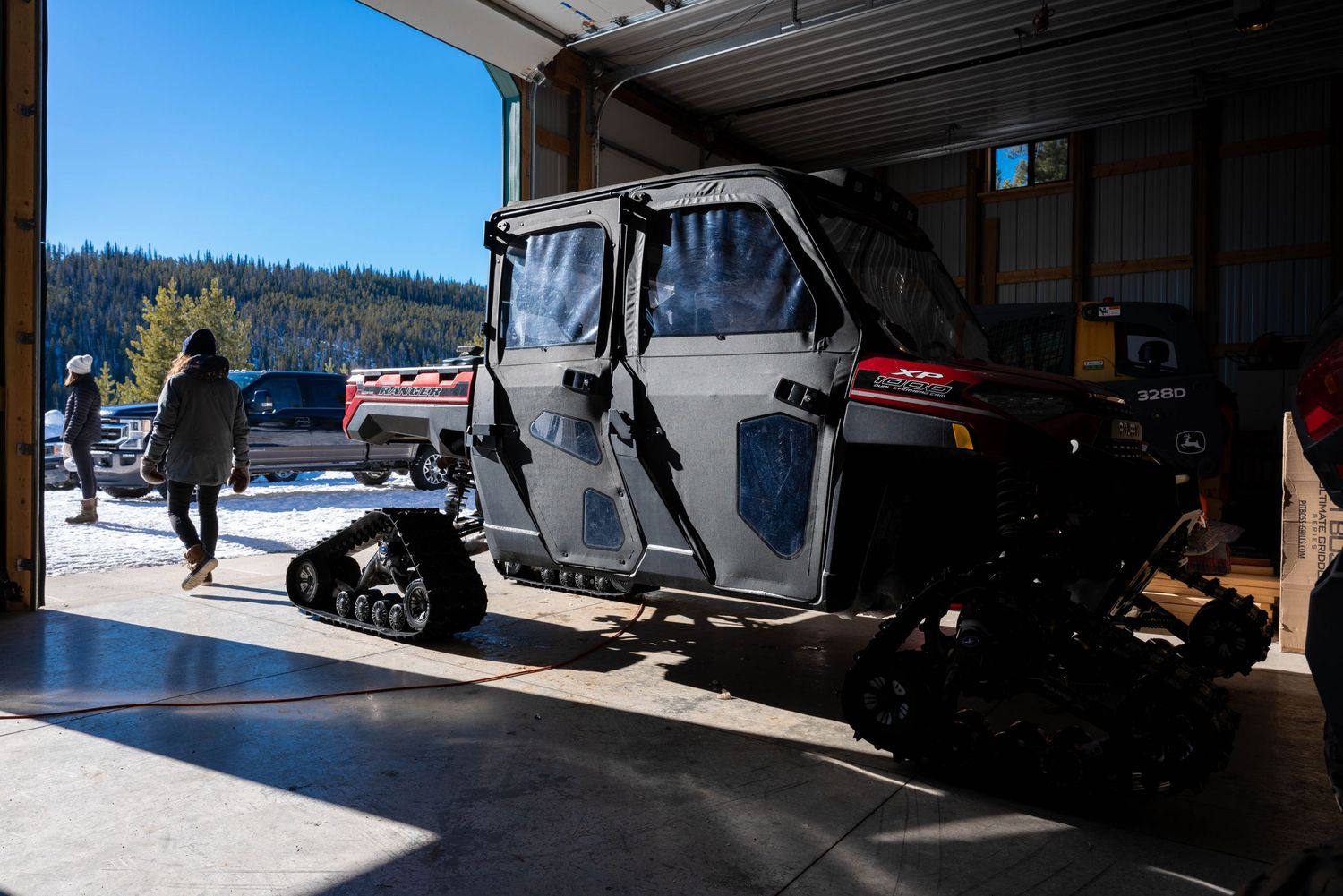 Snow track utility vehicle parked inside a garage with snowy mountain landscape visible outside.