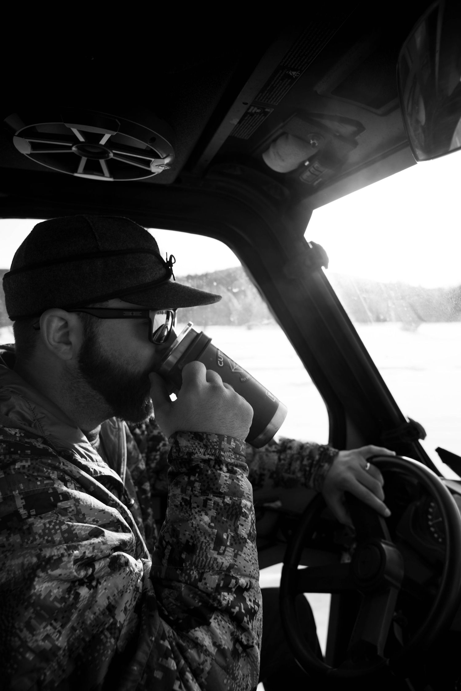 Black and white portrait of a bearded man in camo drinking from a thermos while driving.