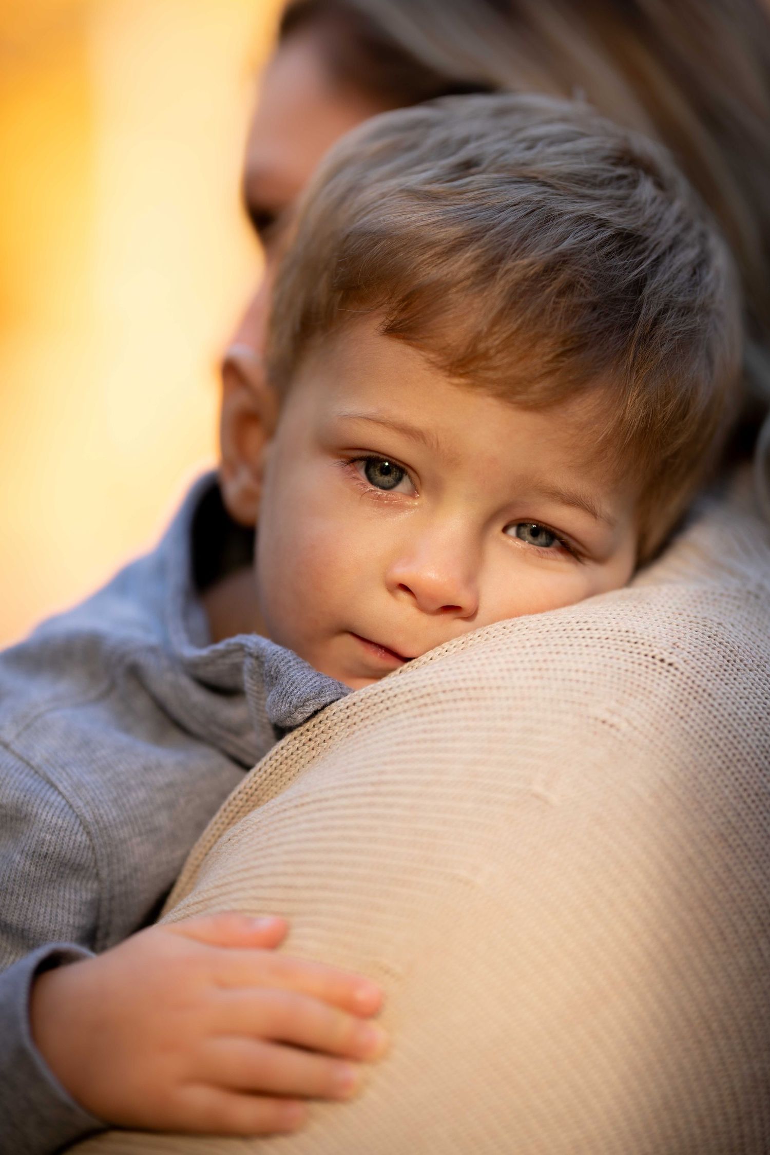 Close-up of a young toddler boy with blue eyes resting his head on a pregnant mother's belly outdoors.
