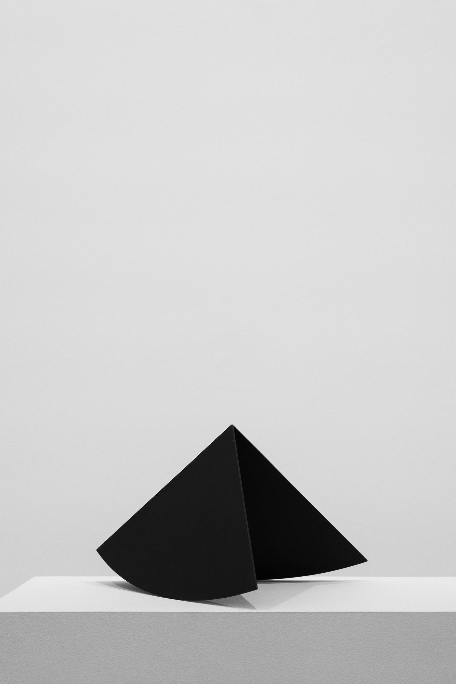 Minimalist black geometric triangular sculpture on white pedestal against gray background.
