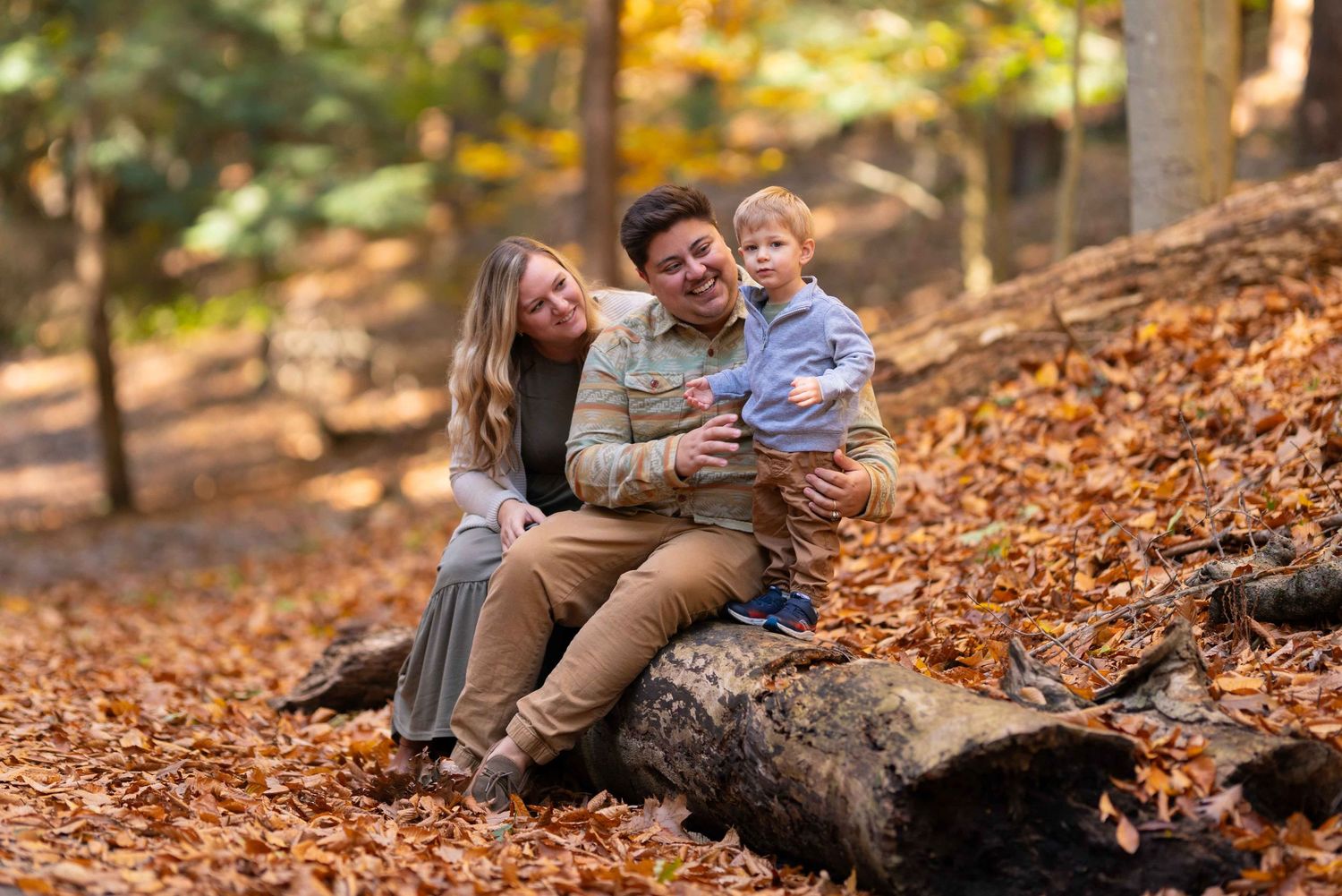 A happy family sits on a log surrounded by fall foliage in a sunlit autumn forest.