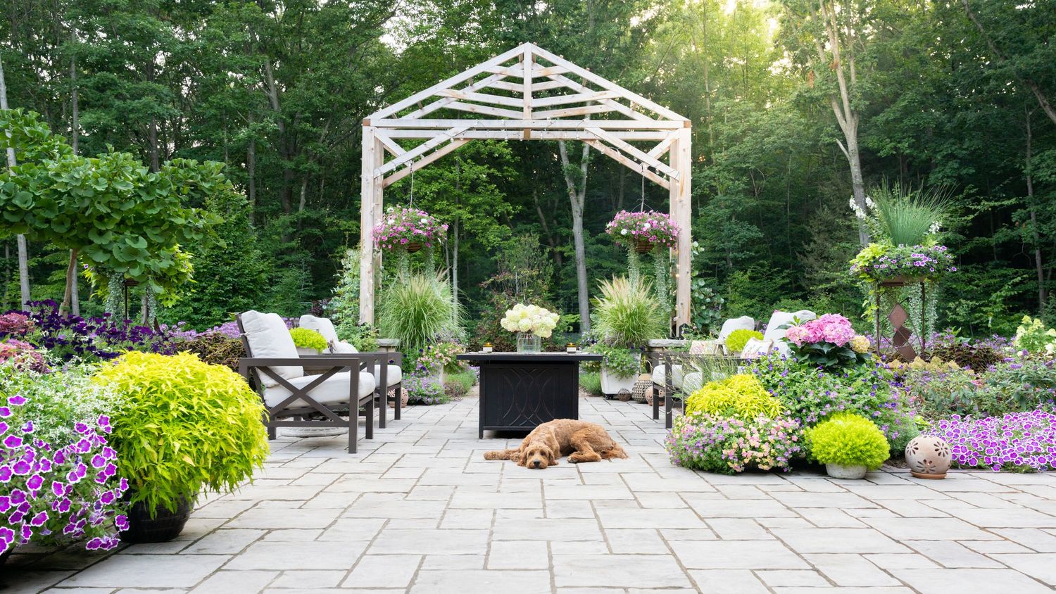 Elegant patio garden with white pergola, fire pit, colorful flowers, and a golden retriever relaxing.