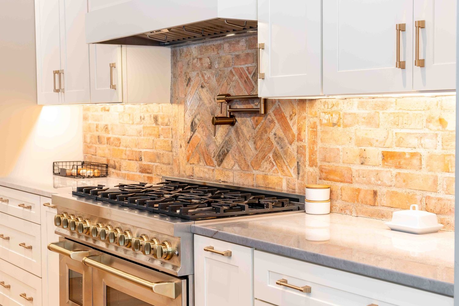 White kitchen with herringbone brick backsplash, gold hardware, and stainless steel range.