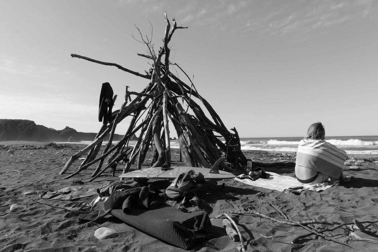 Person sitting beside a driftwood teepee structure on a black and white beach scene.