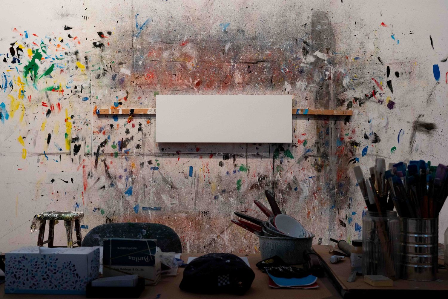 Artist studio wall splattered with colorful paint surrounding a blank white canvas.