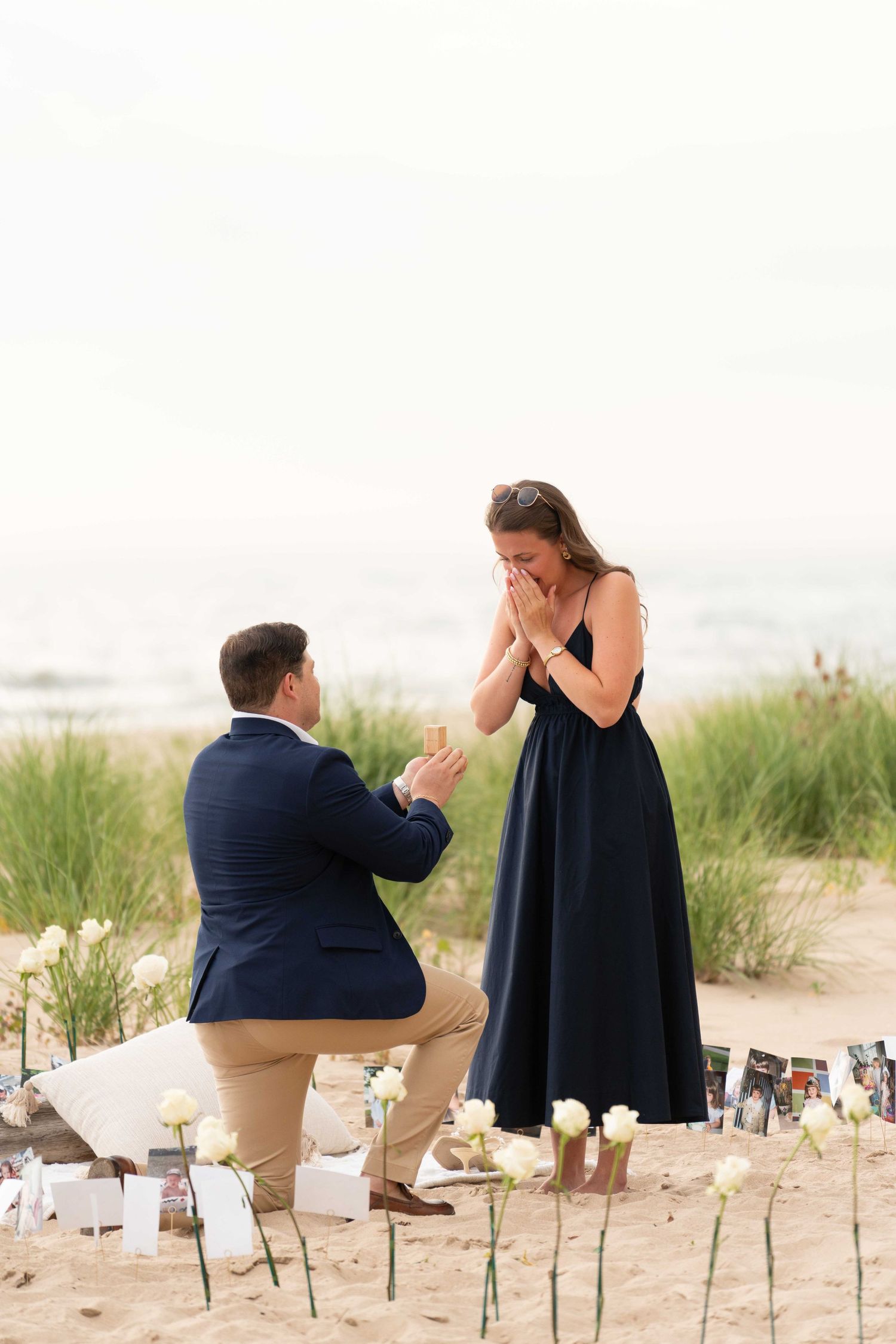 A man proposes on a sandy beach with white roses as a woman in a black dress reacts with surprise.