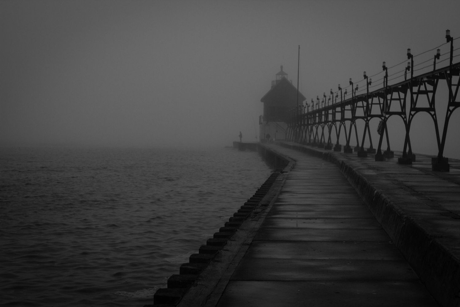 Black and white foggy pier leading to a lighthouse over calm water, creating a moody and mysterious atmosphere.