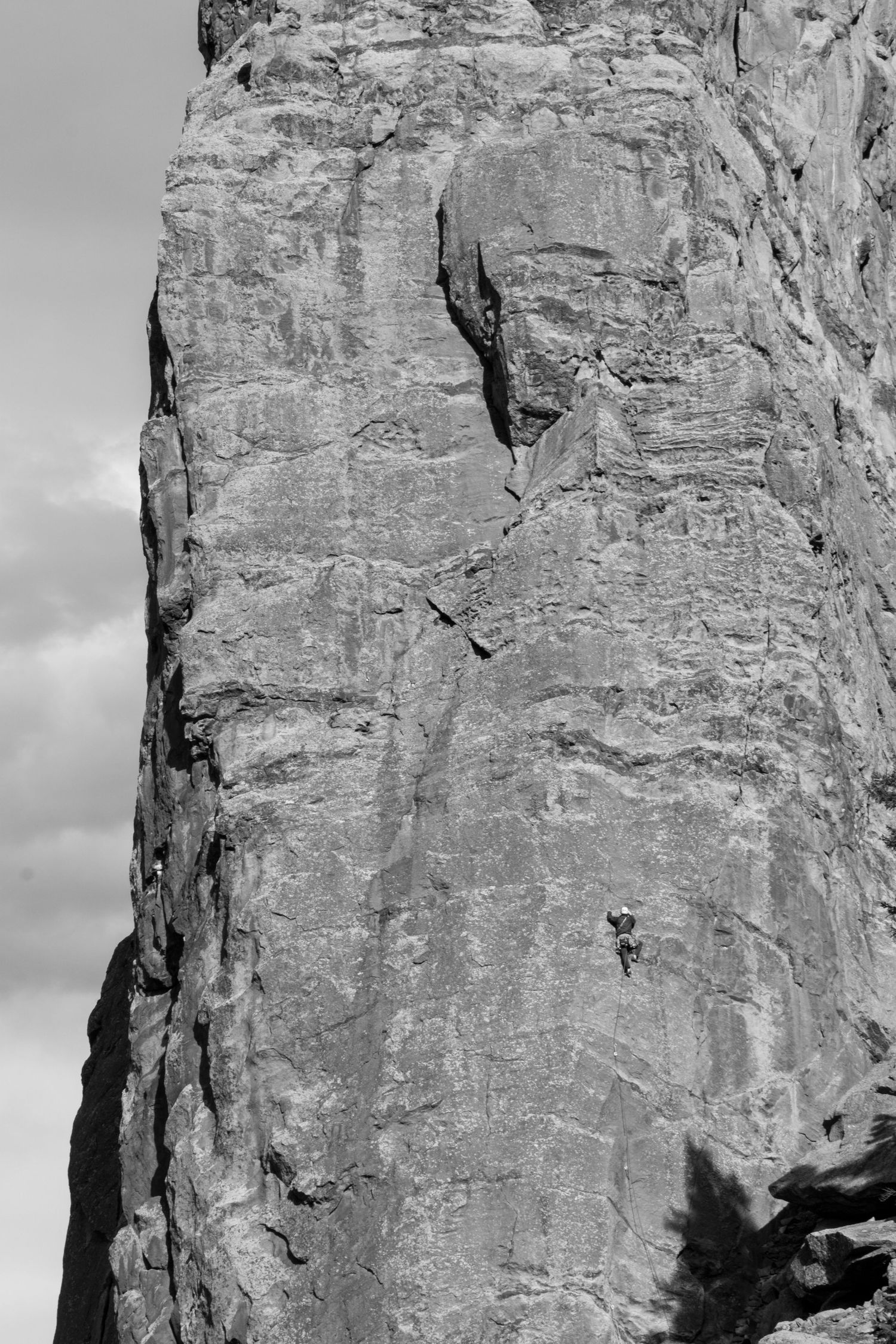 Lone climber ascends a massive vertical rock face in dramatic black and white photo.