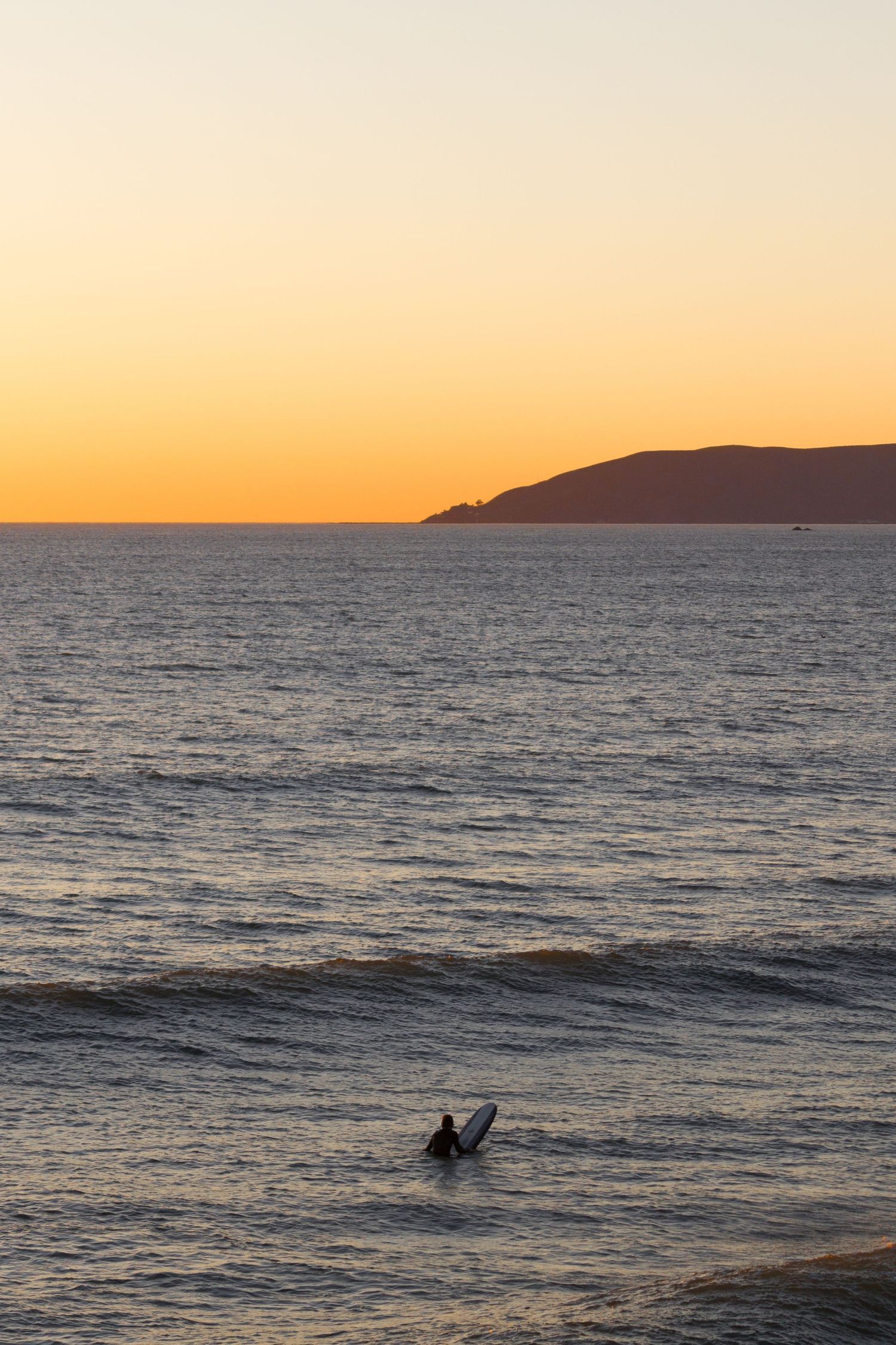 Lone surfer paddling out at sunset with golden sky and coastal headland in background.