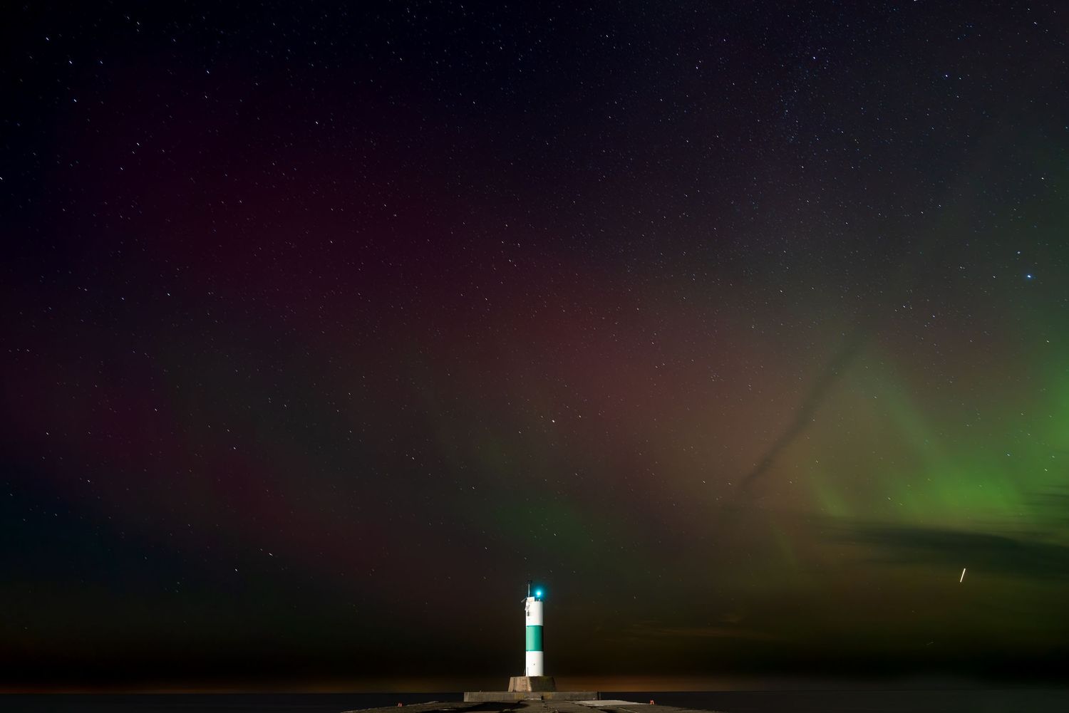 Lone lighthouse standing on snowy pier beneath colorful green and purple northern lights aurora display.