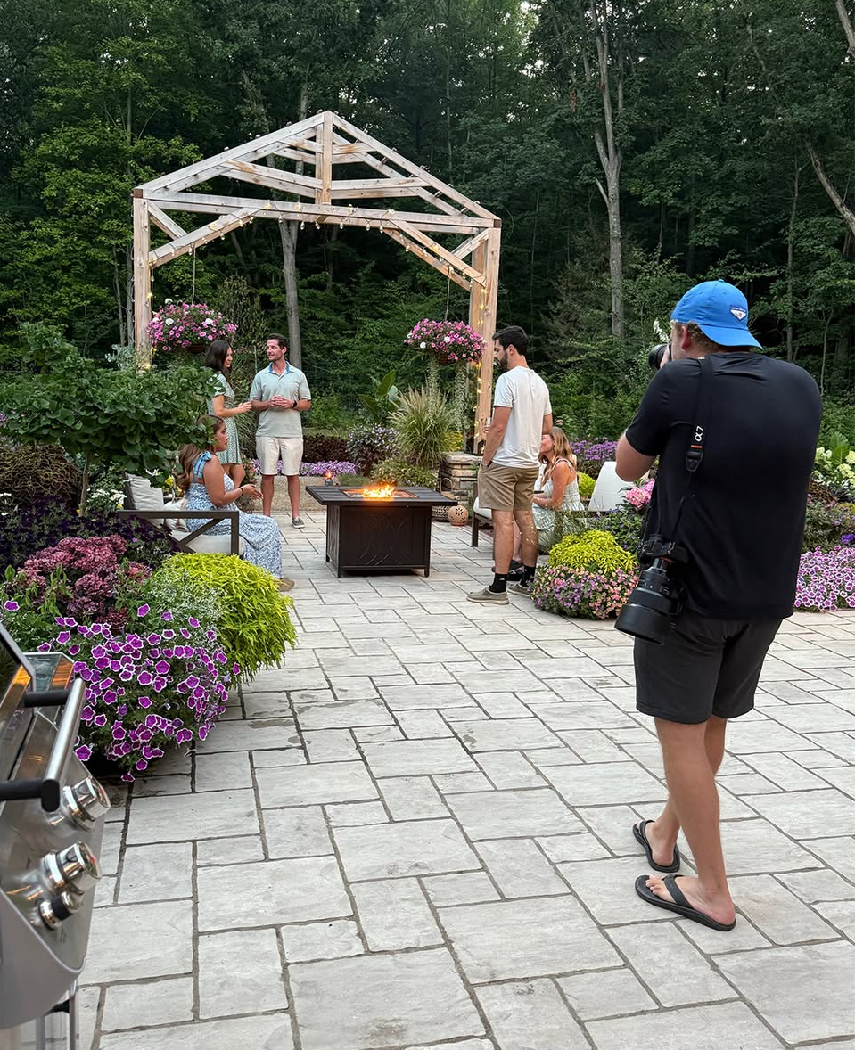 Photographer in a blue cap shooting subjects near a wooden gazebo surrounded by colorful flowers.