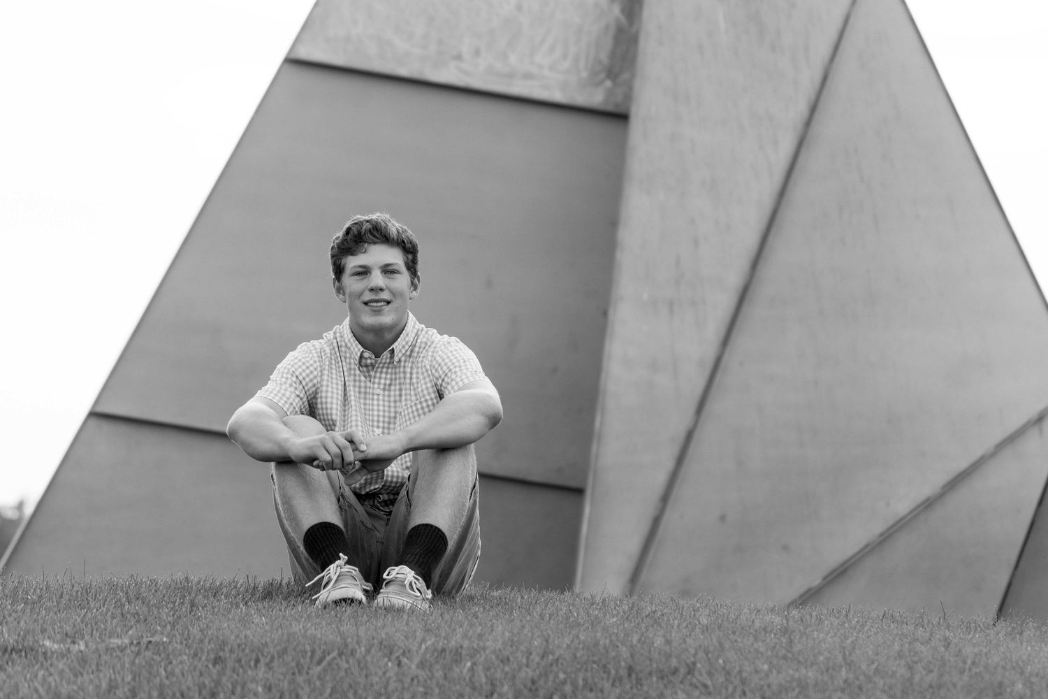 A young man sits cross-legged on grass in front of large geometric concrete structures in black and white.