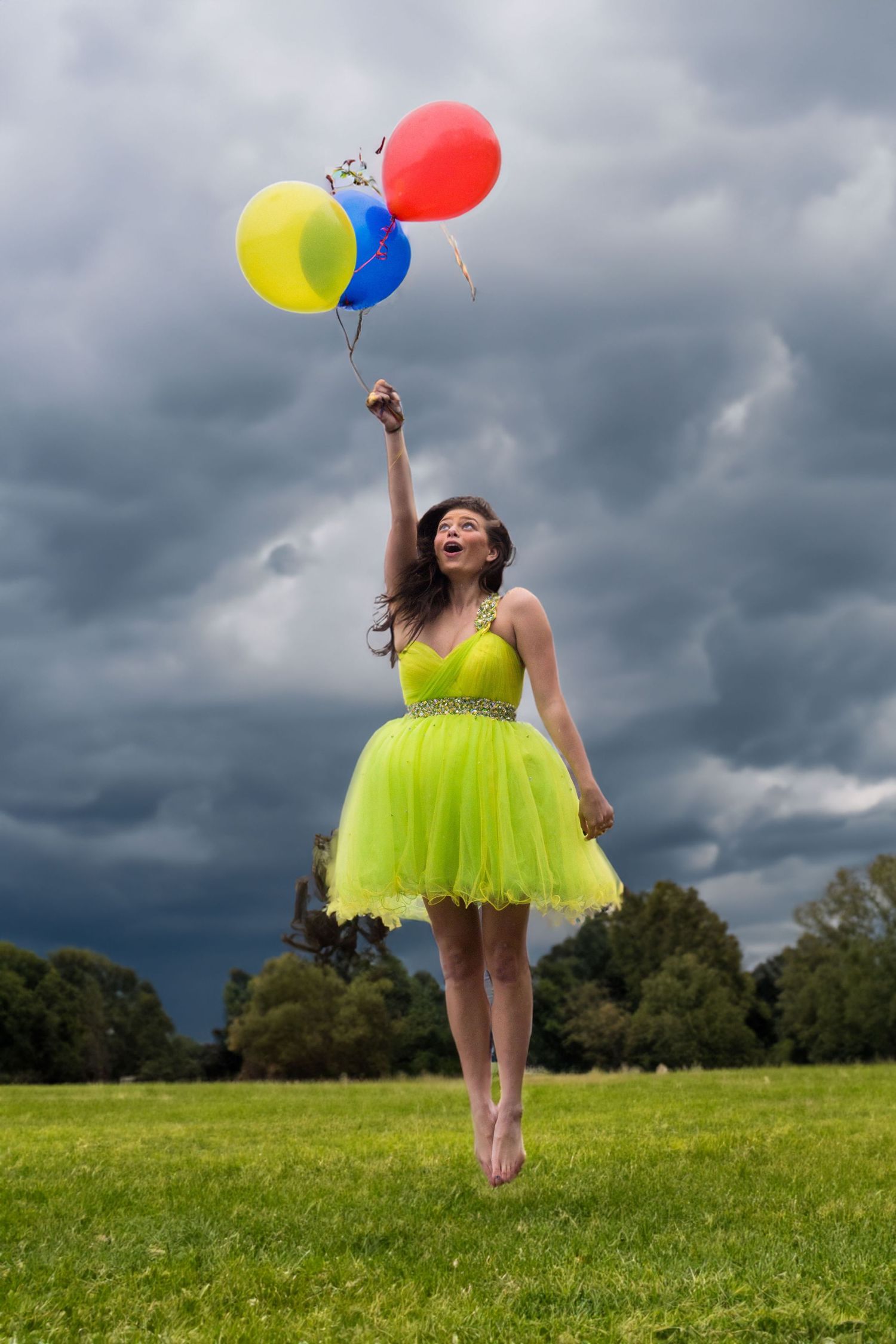 A young woman in a yellow tutu dress jumps while holding colorful balloons under a stormy sky.