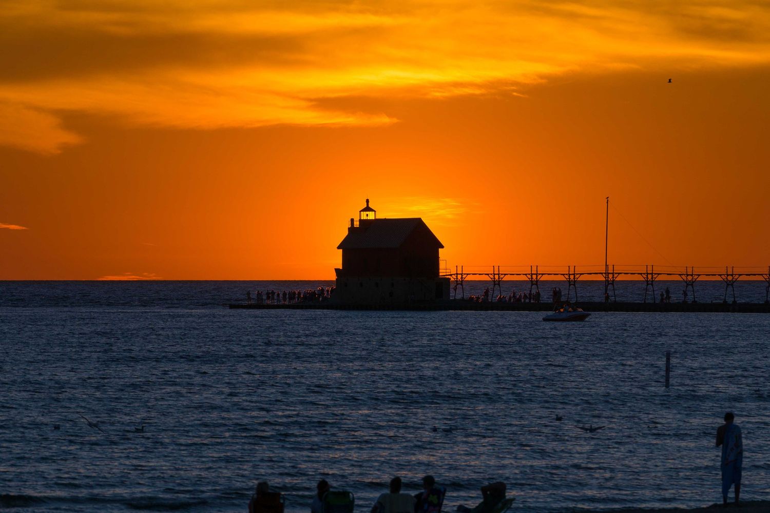 Silhouette of a lighthouse and pier at sunset with people gathered on a beach in warm orange light.