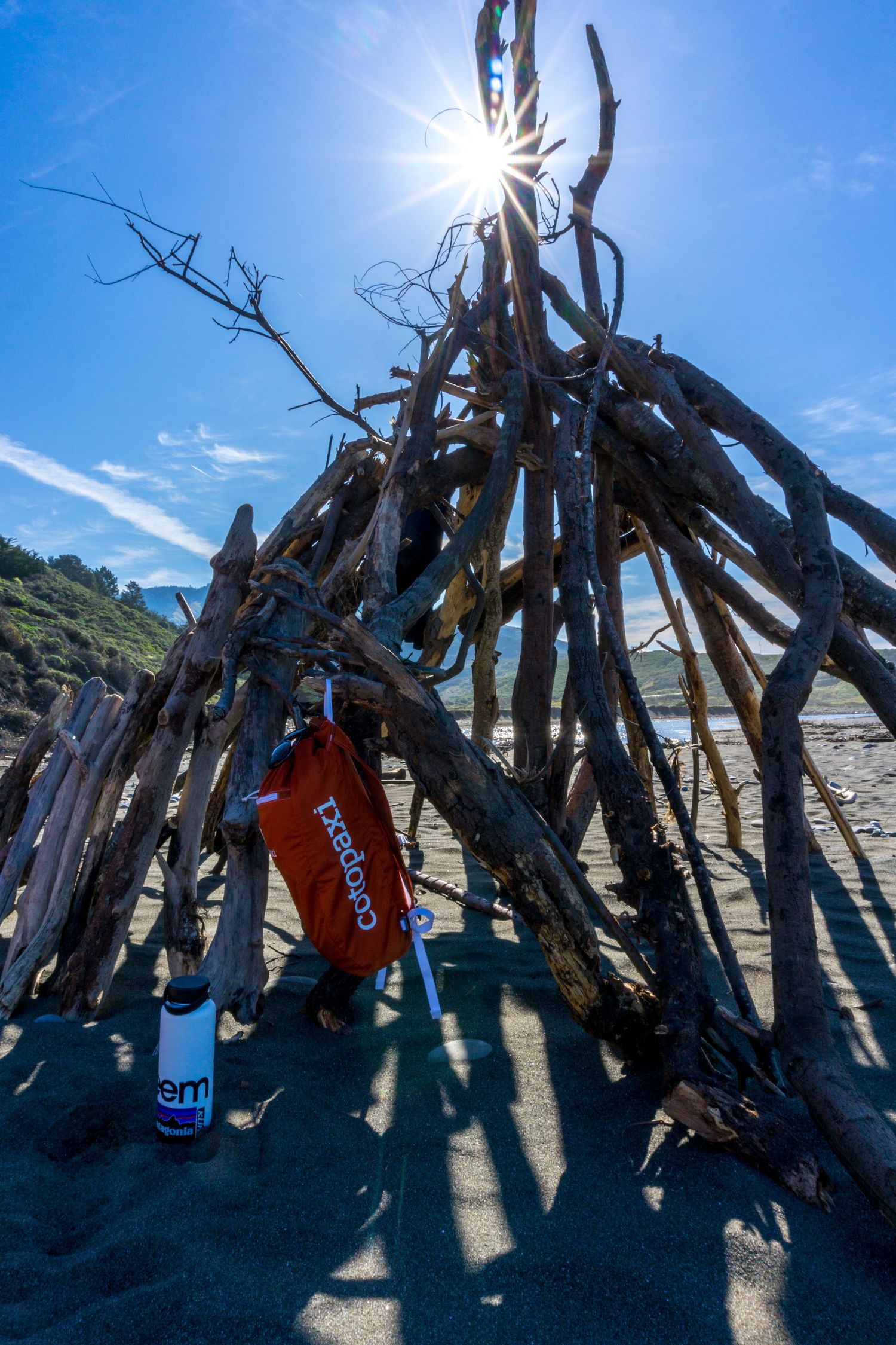 Driftwood teepee structure on black sand beach with sun flare and Colgate products in foreground.