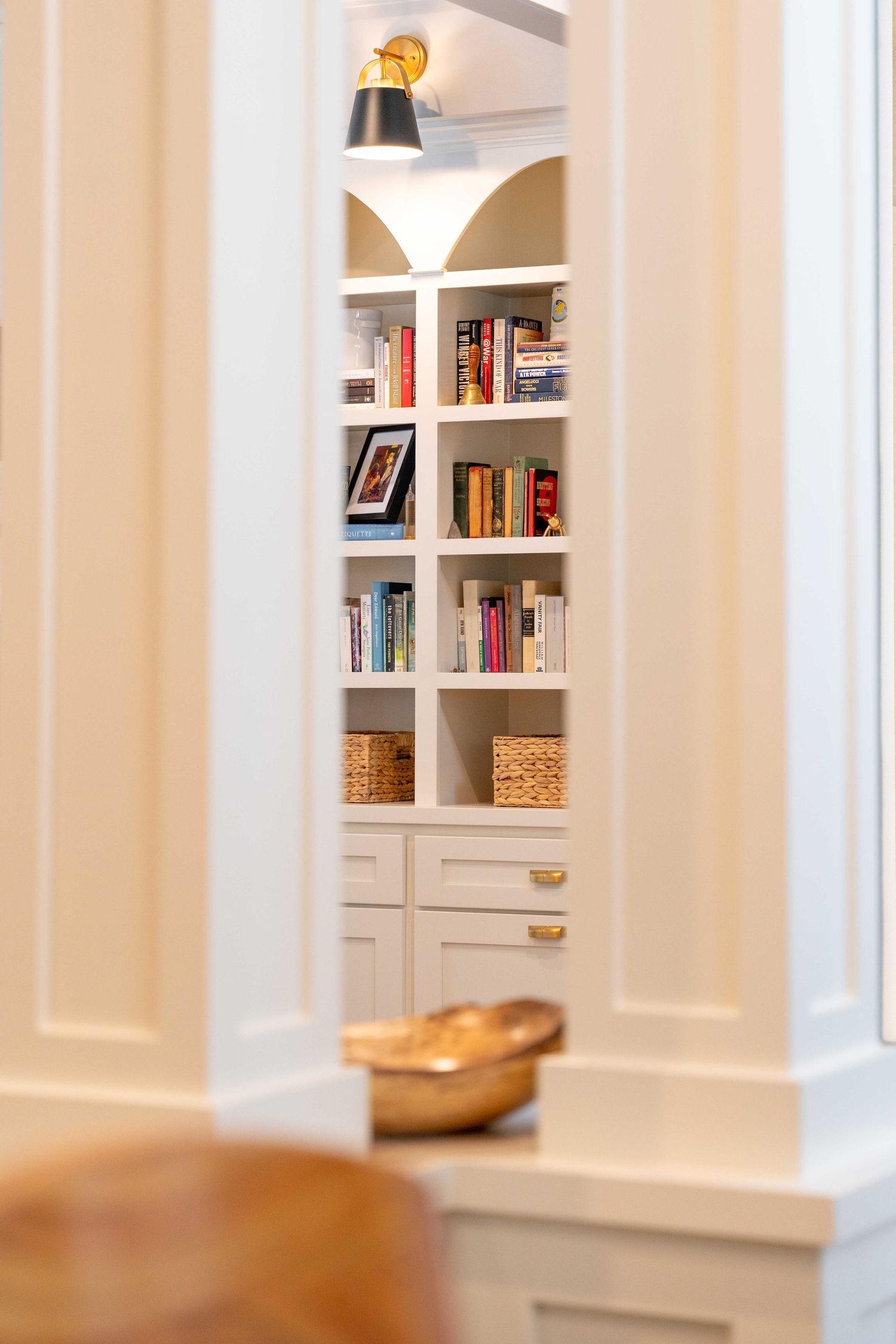 Built-in white bookshelf with books, wicker baskets, and brass hardware accents in a home library.
