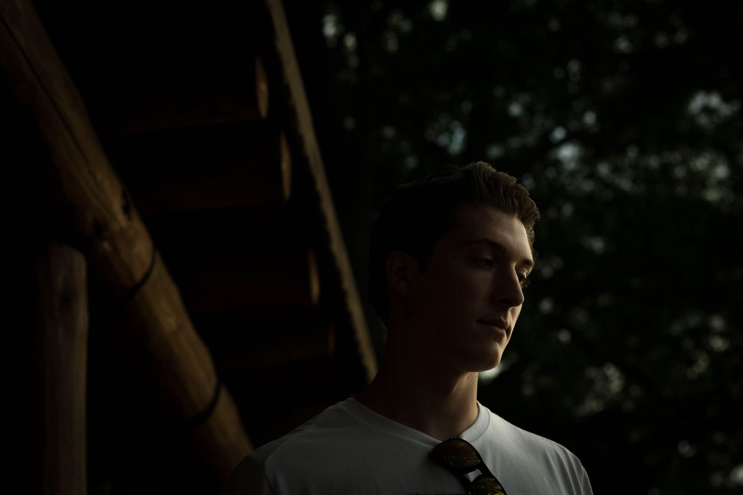 A young man in a white shirt and tie stands in dramatic shadow near a wooden structure.