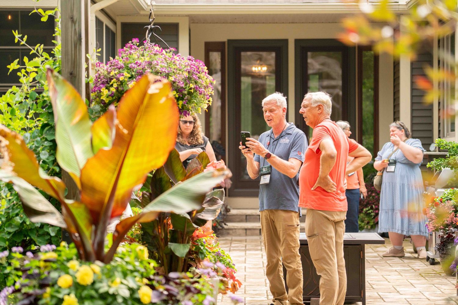 People socializing in a beautifully landscaped garden patio with colorful hanging flowers and tropical plants.