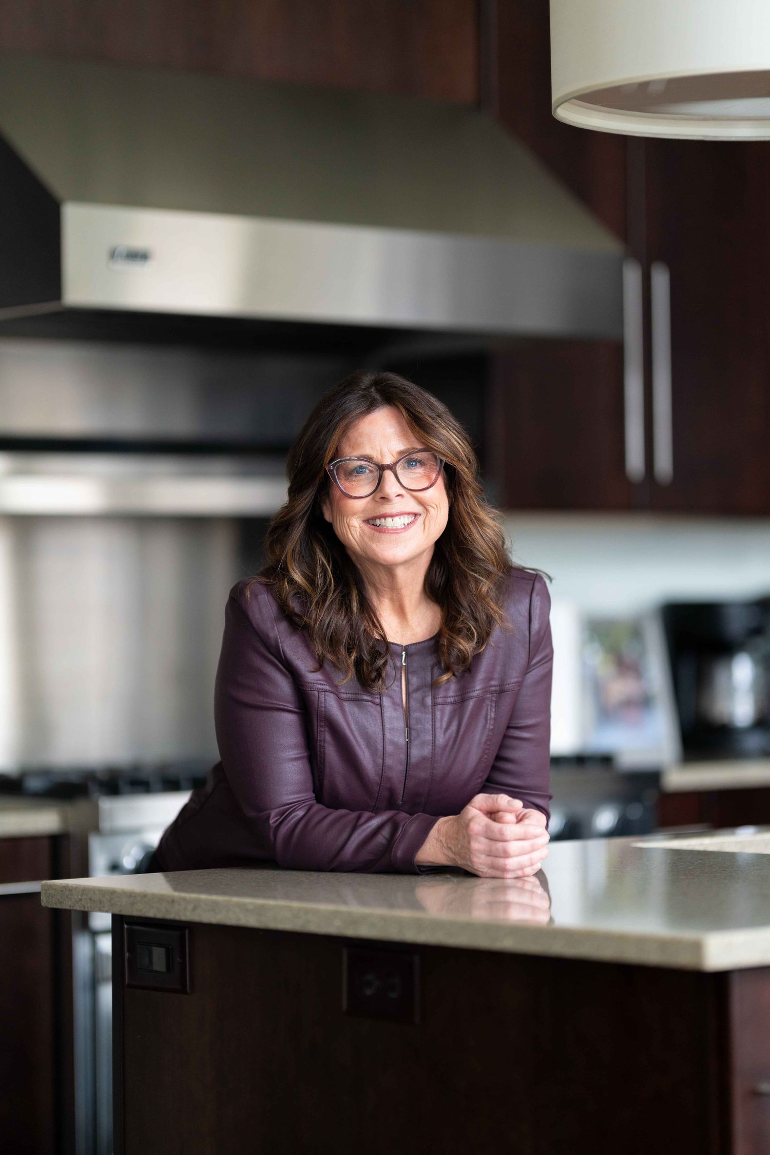 A smiling middle-aged woman with glasses leans on a kitchen island in a purple leather jacket.