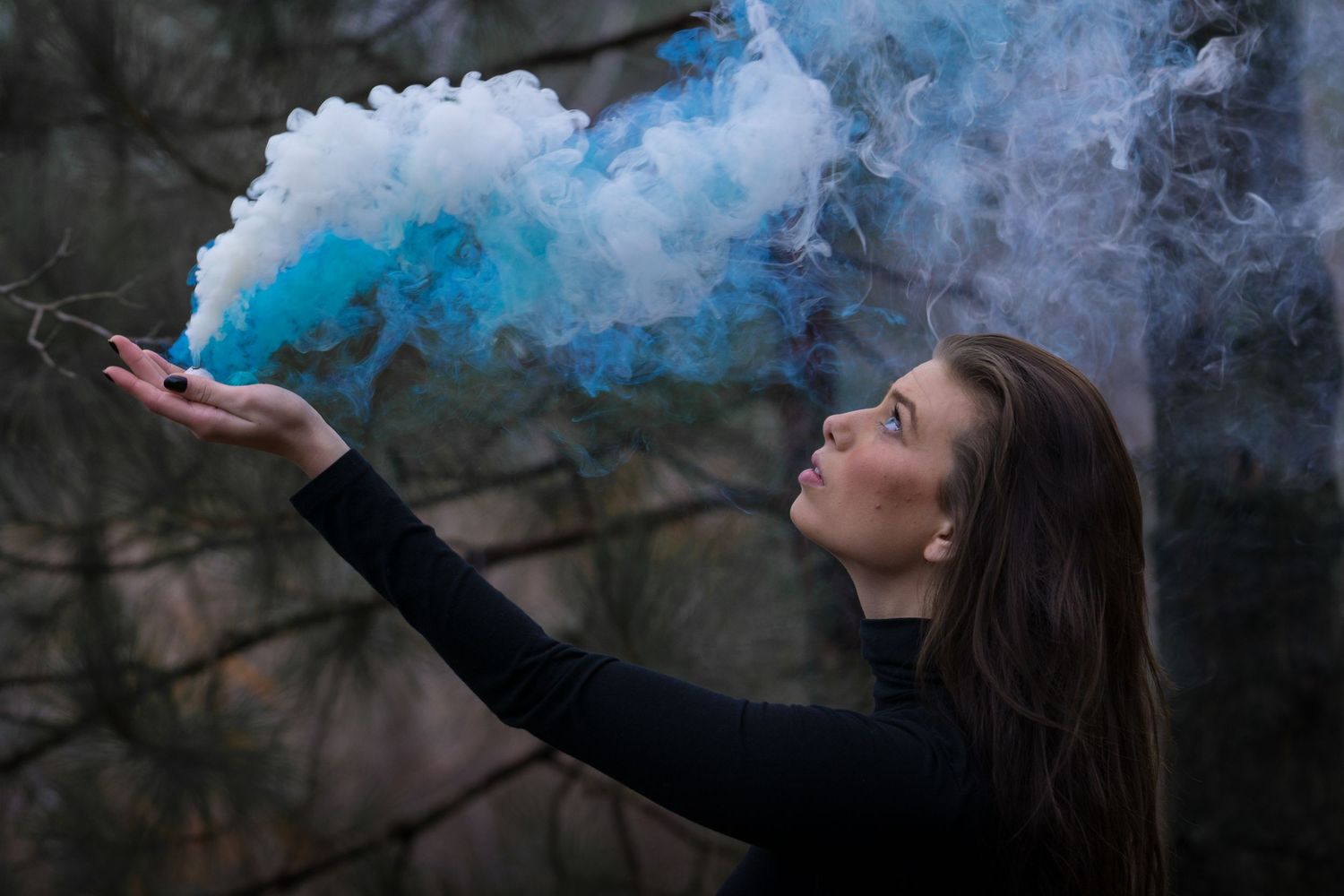 A young woman in black blows colorful blue and white smoke from her outstretched hand outdoors.
