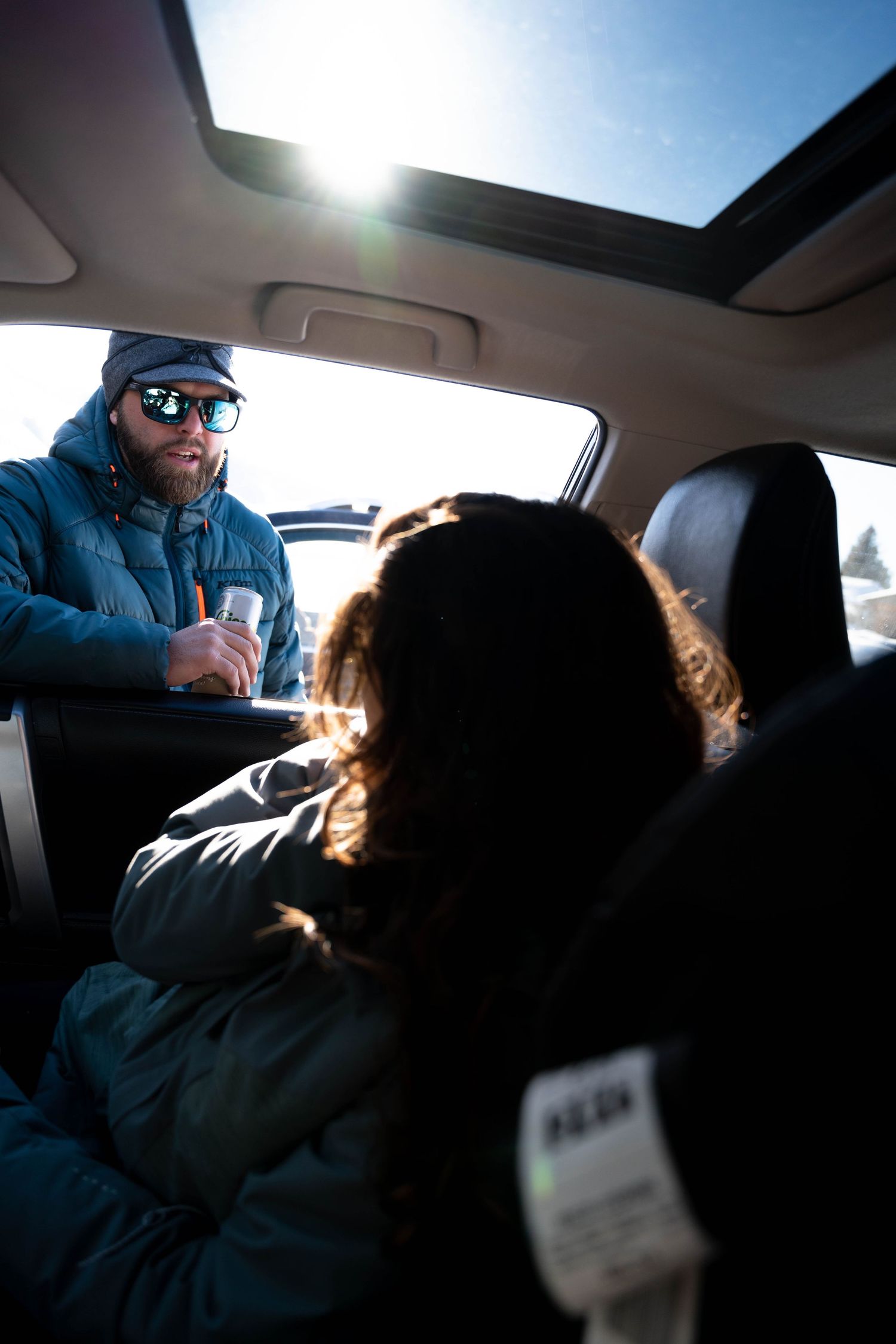 Bearded man in winter gear holding a drink speaking to passengers inside a sunlit vehicle.