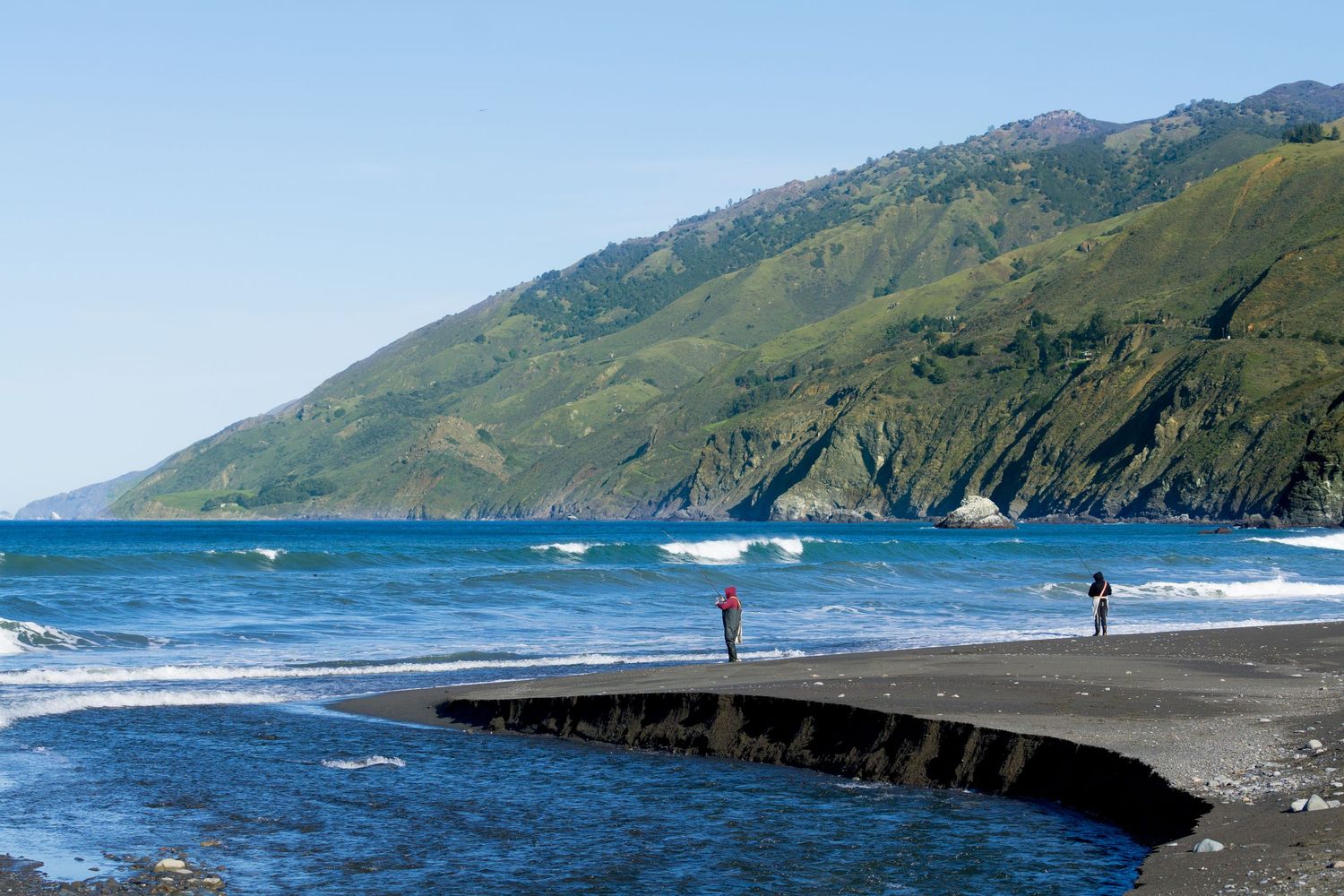 Two people standing on black sand beach with dramatic green coastal cliffs and blue ocean waves.