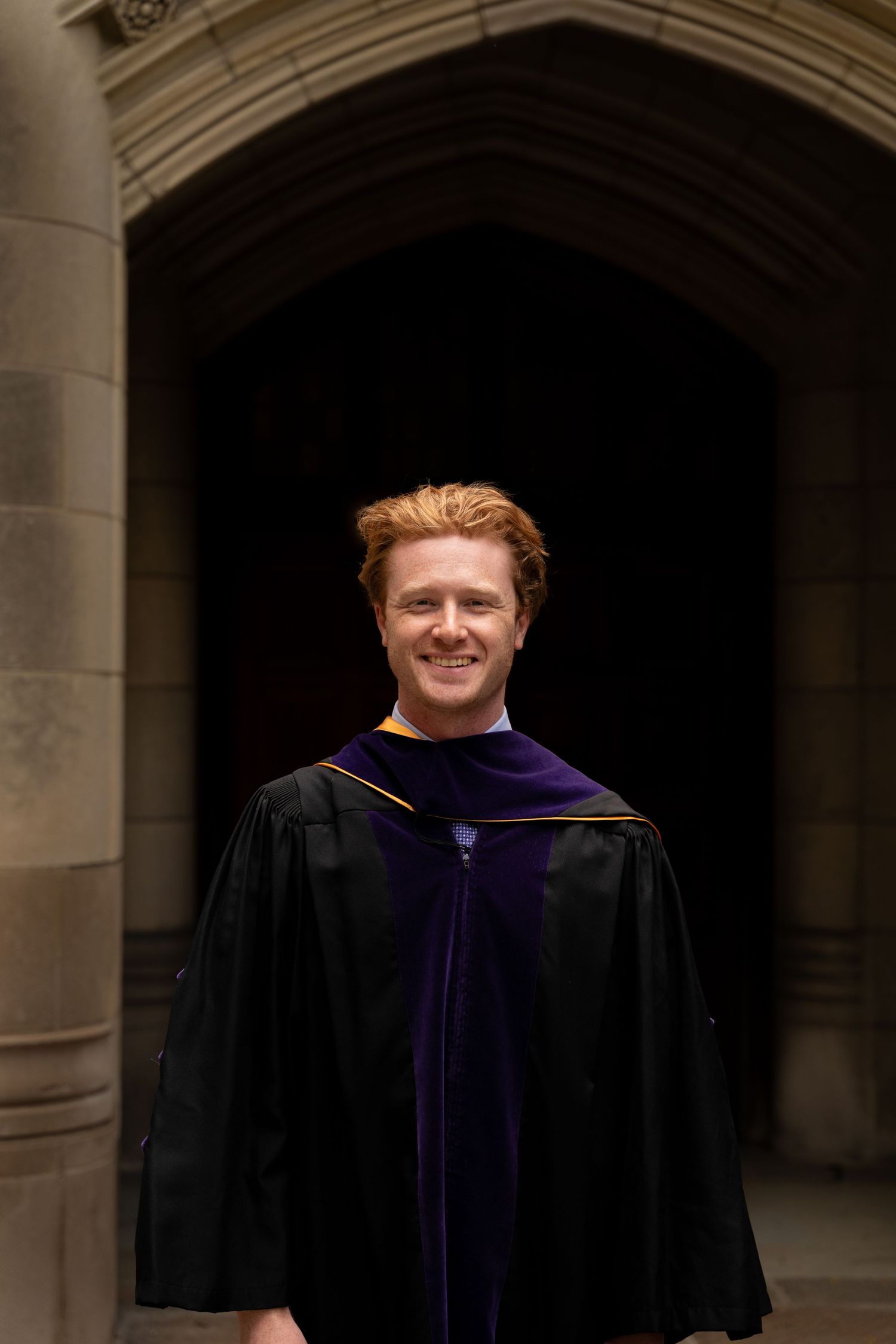 Smiling graduate in black and purple law school regalia standing under stone archway.