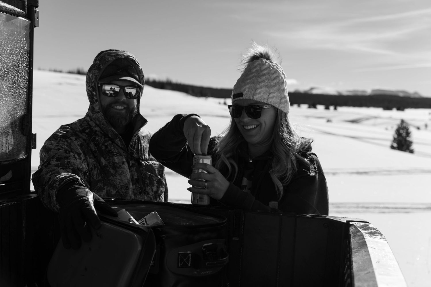 Black and white photo of a smiling couple holding drinks outdoors beside an off-road vehicle in snow.