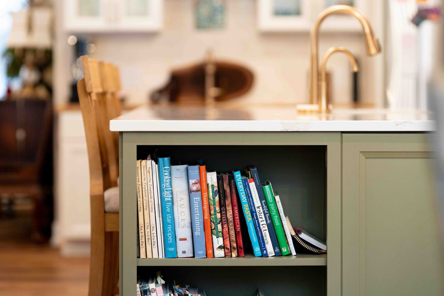 Green kitchen island shelf stocked with colorful cookbooks near a gold faucet and white countertop.