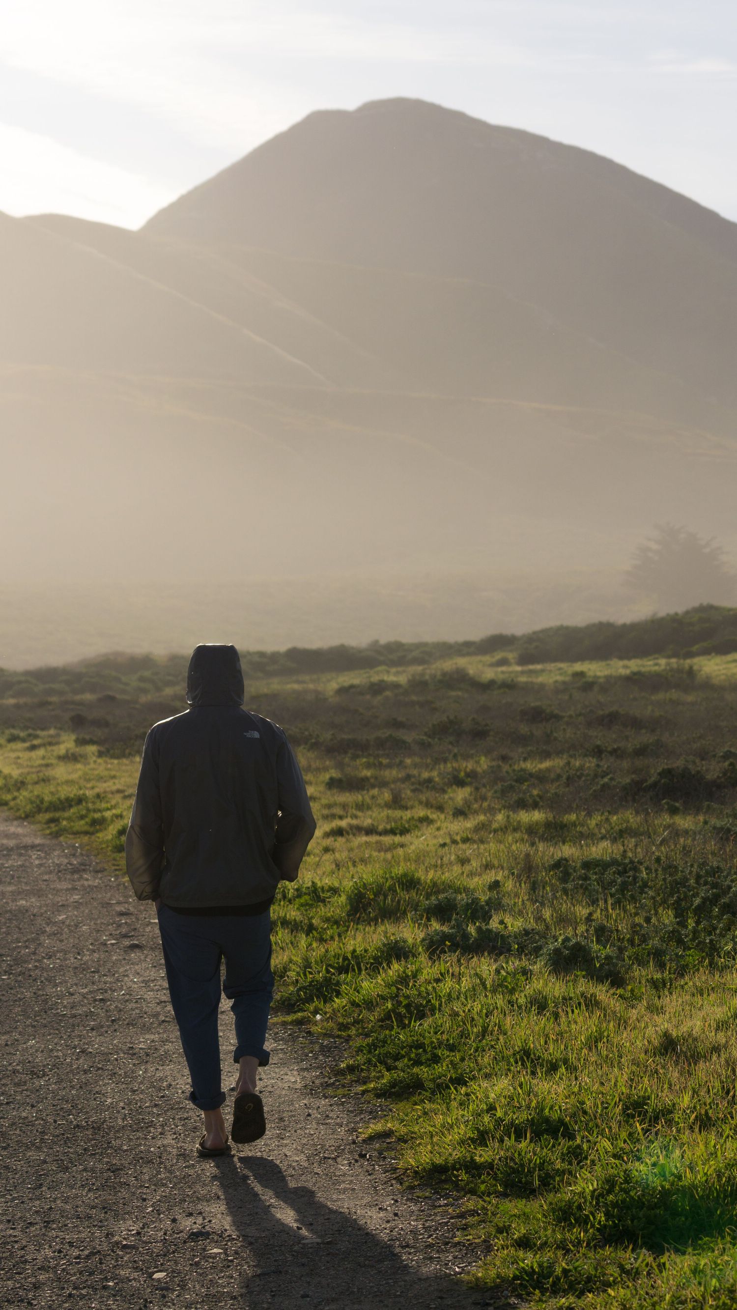 Person in North Face jacket walking misty trail toward mountain peak in golden morning light.