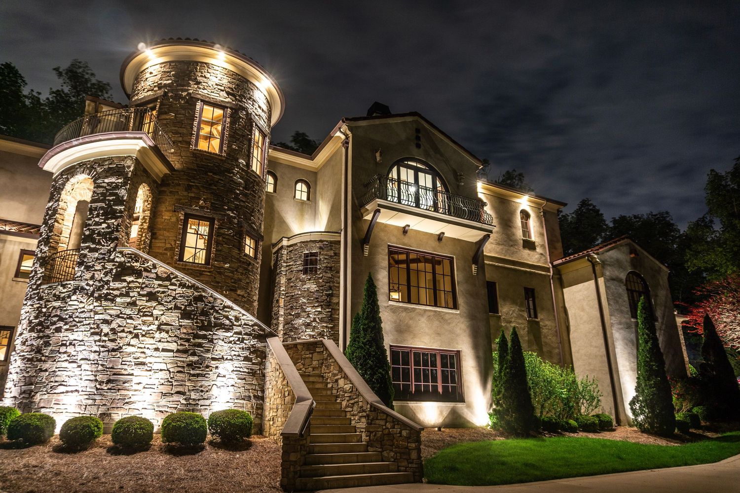 Illuminated luxury stone and stucco castle-style home exterior at night with landscape lighting.