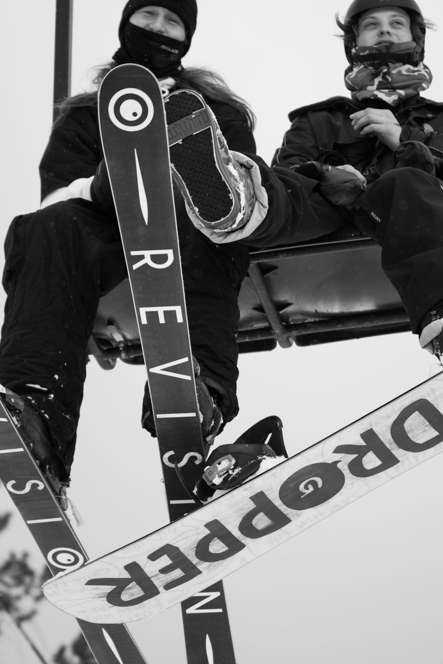 Two snowboarders ride a ski lift with Revision and Dropper branded snowboards visible.