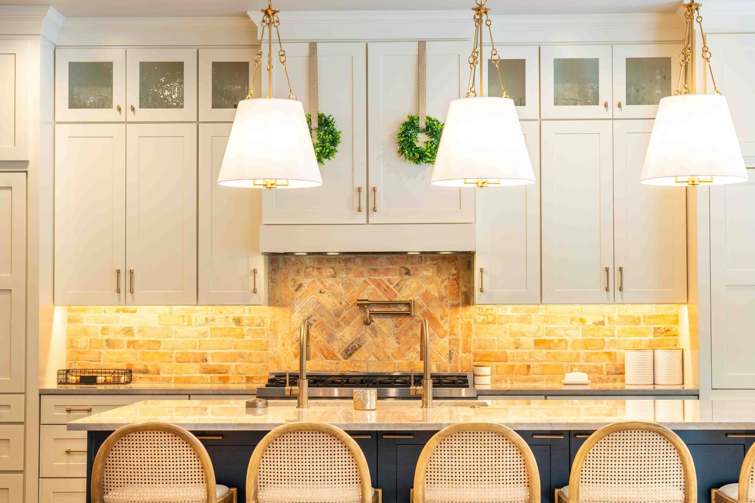 Luxury kitchen with brick backsplash, white pendant lights, rattan bar stools, and sage green island.