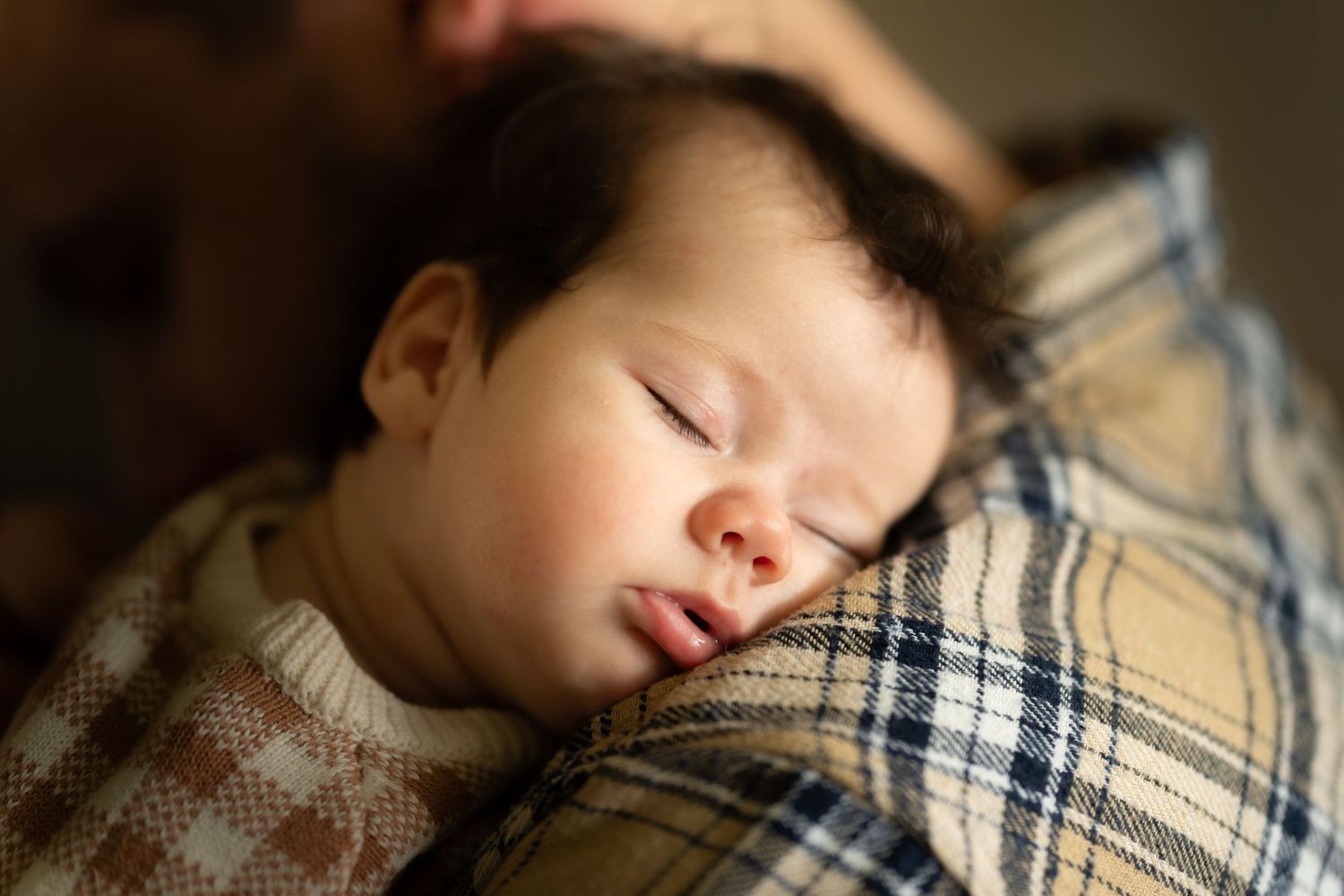 A sleeping newborn baby with dark hair rests peacefully on a plaid blanket on someone's shoulder.