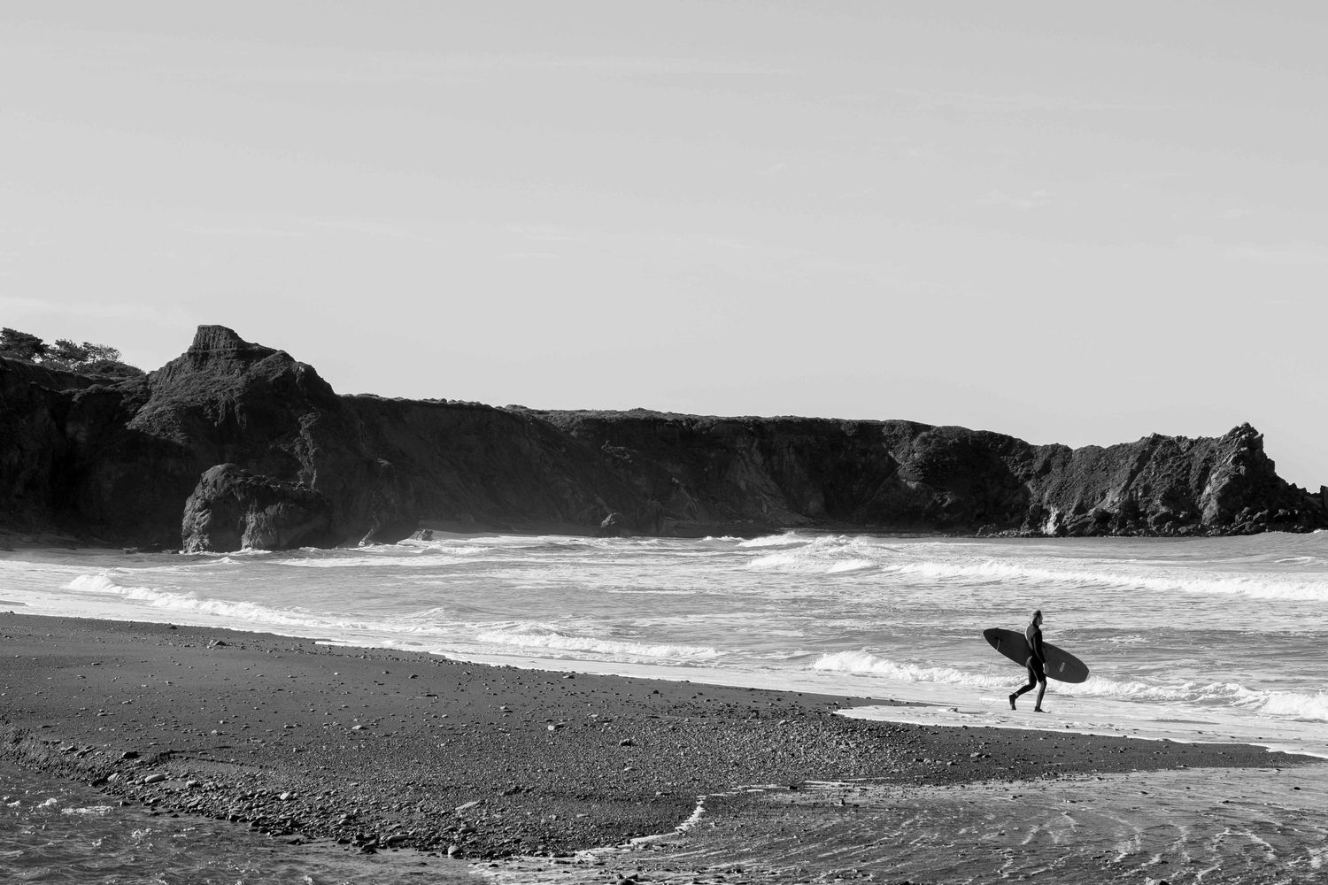 Surfer carrying surfboard along a rocky black and white coastal beach.