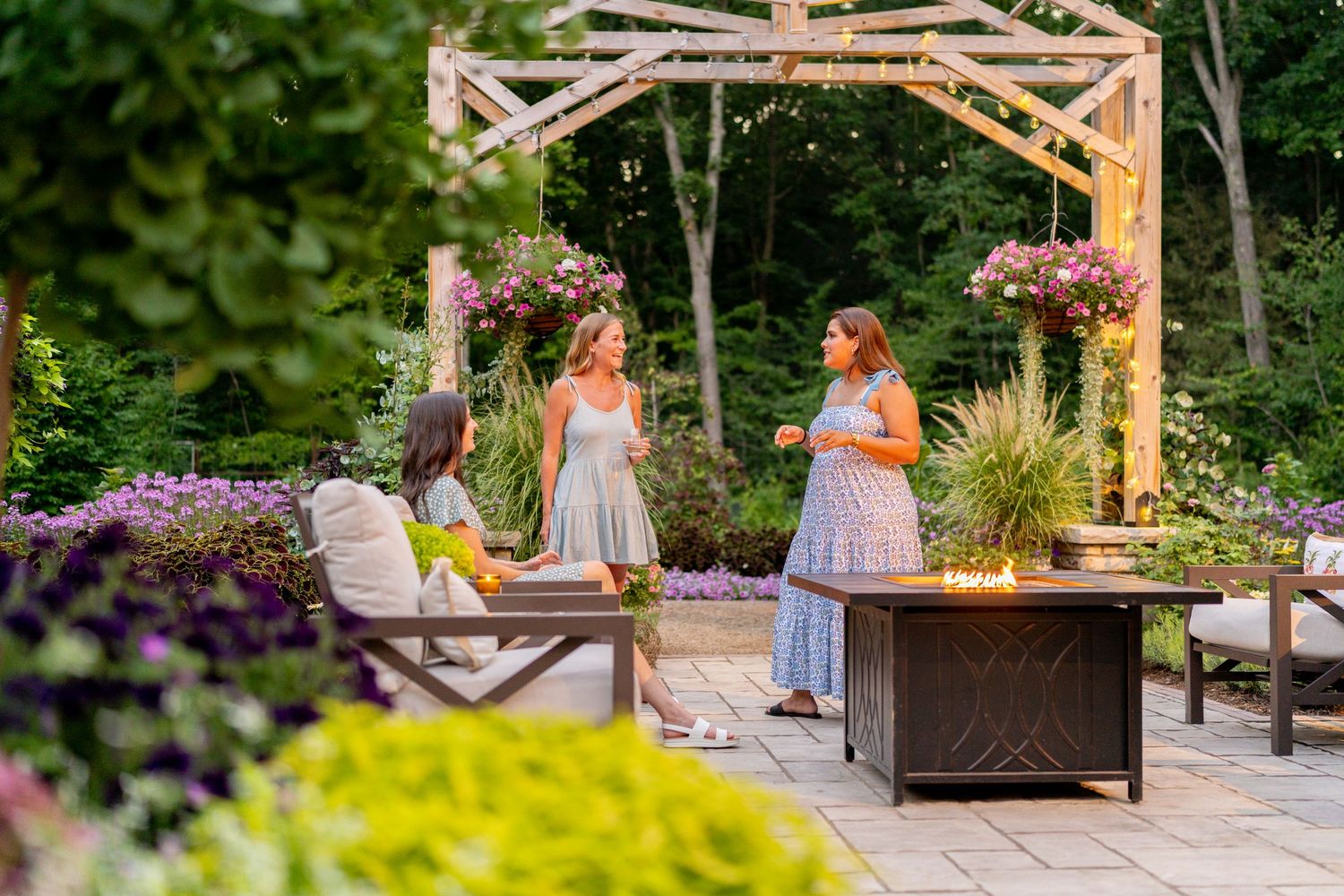 Three women socializing around an outdoor fire pit in a beautifully landscaped garden at dusk.