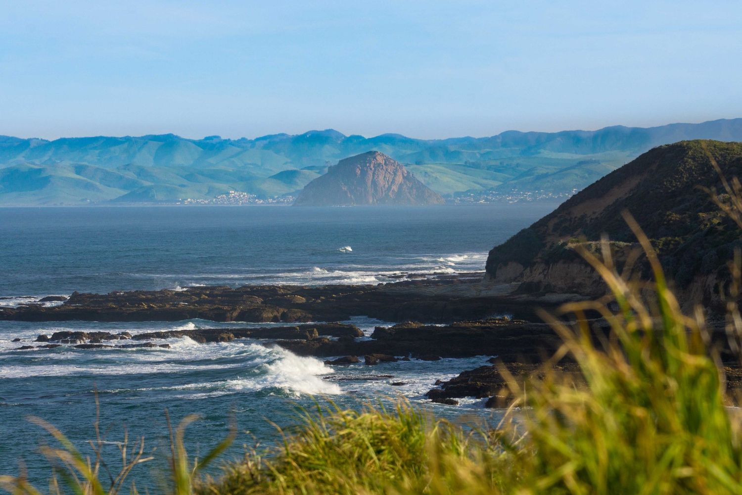 Scenic coastal view with rocky shoreline, ocean waves, and misty blue mountains in the background under a clear sky.