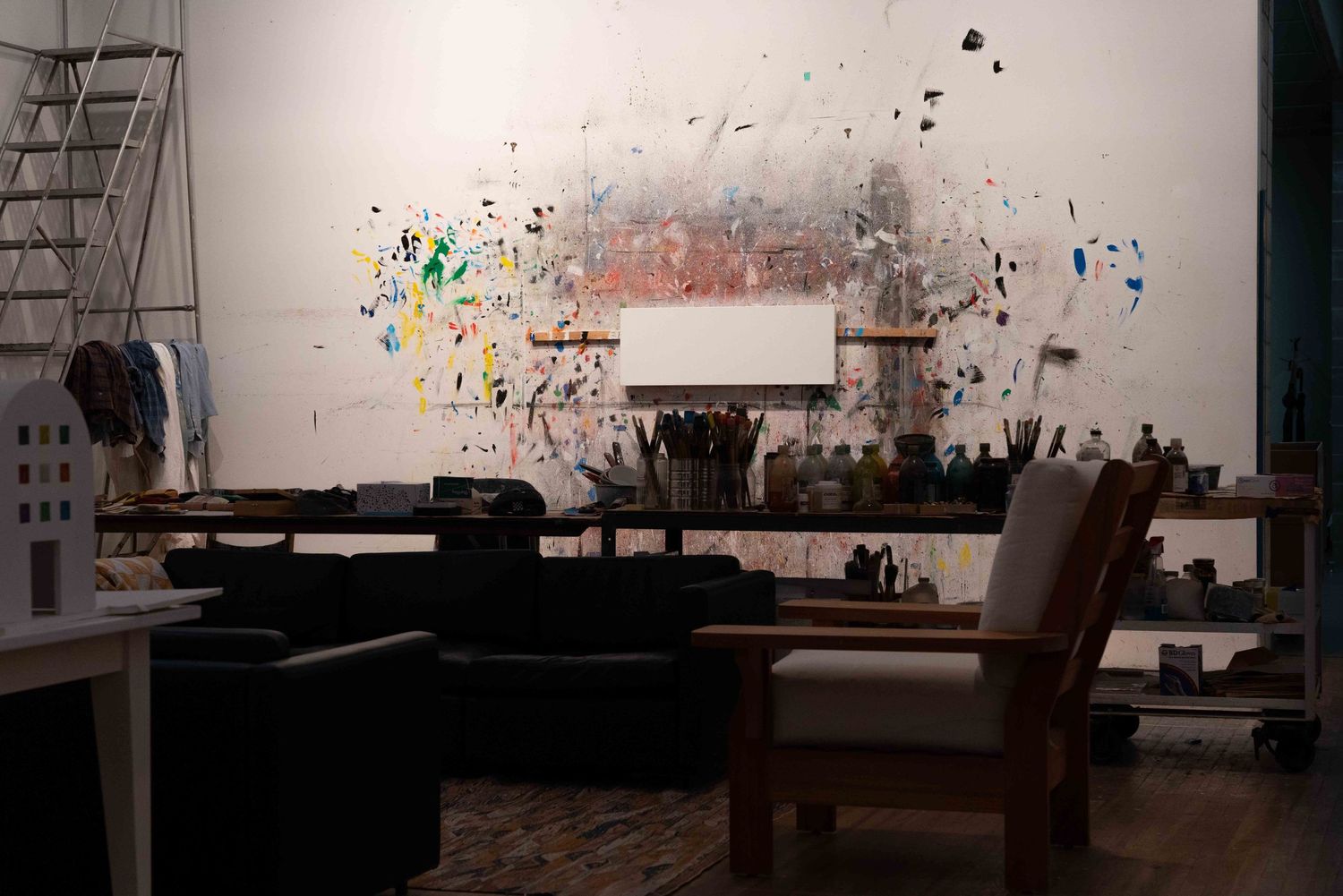 Spacious art studio with paint-splattered wall, blank canvas, brushes, and wooden chair.