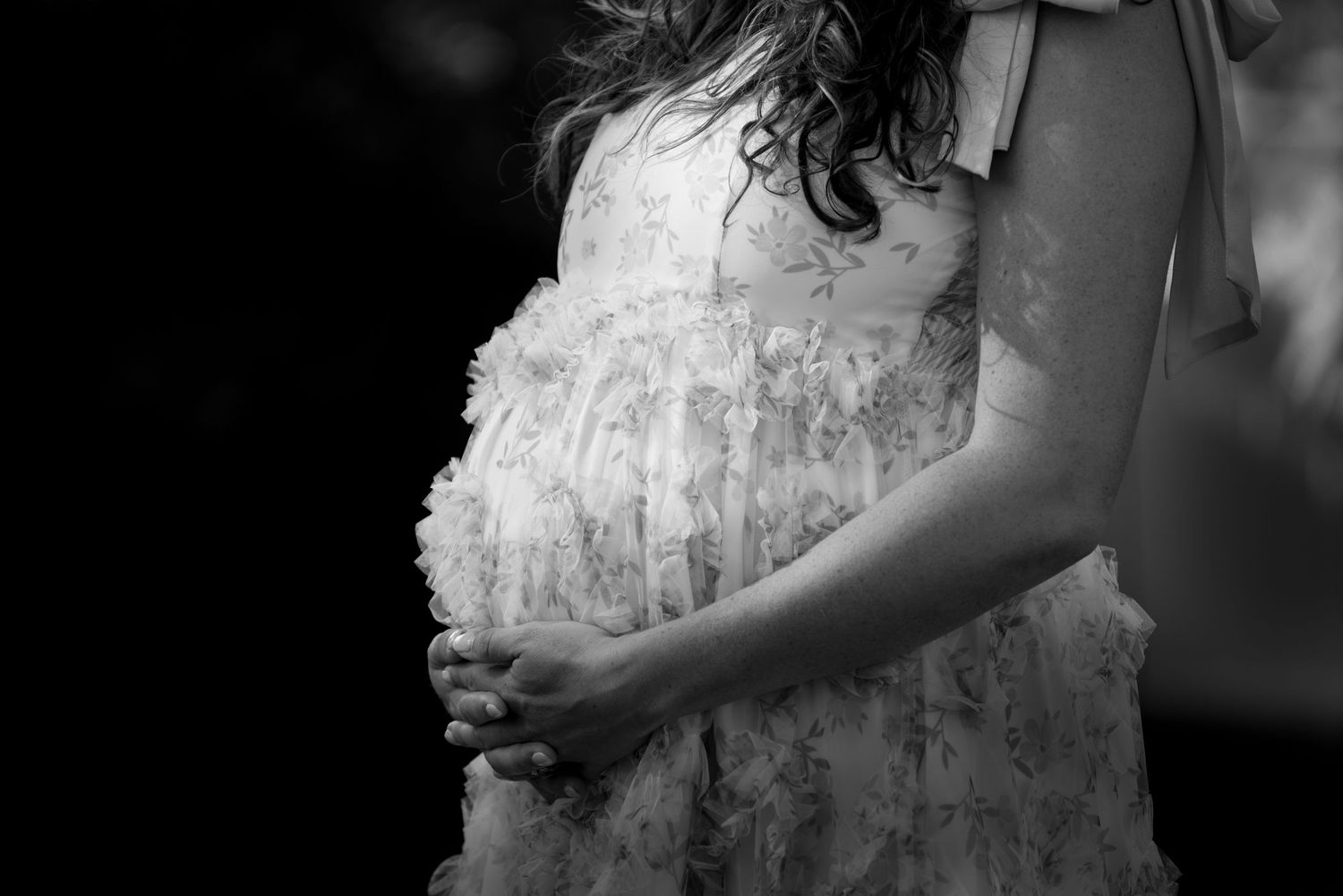 Black and white photo of pregnant woman in floral dress cradling her baby bump.