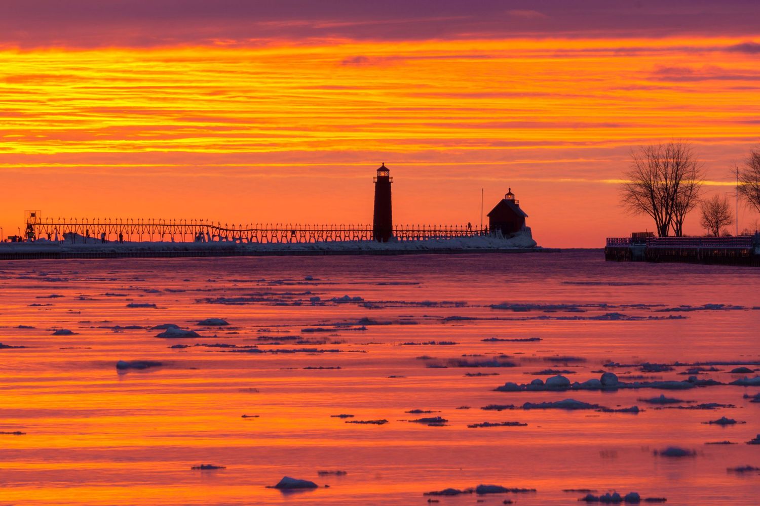 Dramatic fiery orange sunset silhouettes a lighthouse and pier over icy waters in winter.