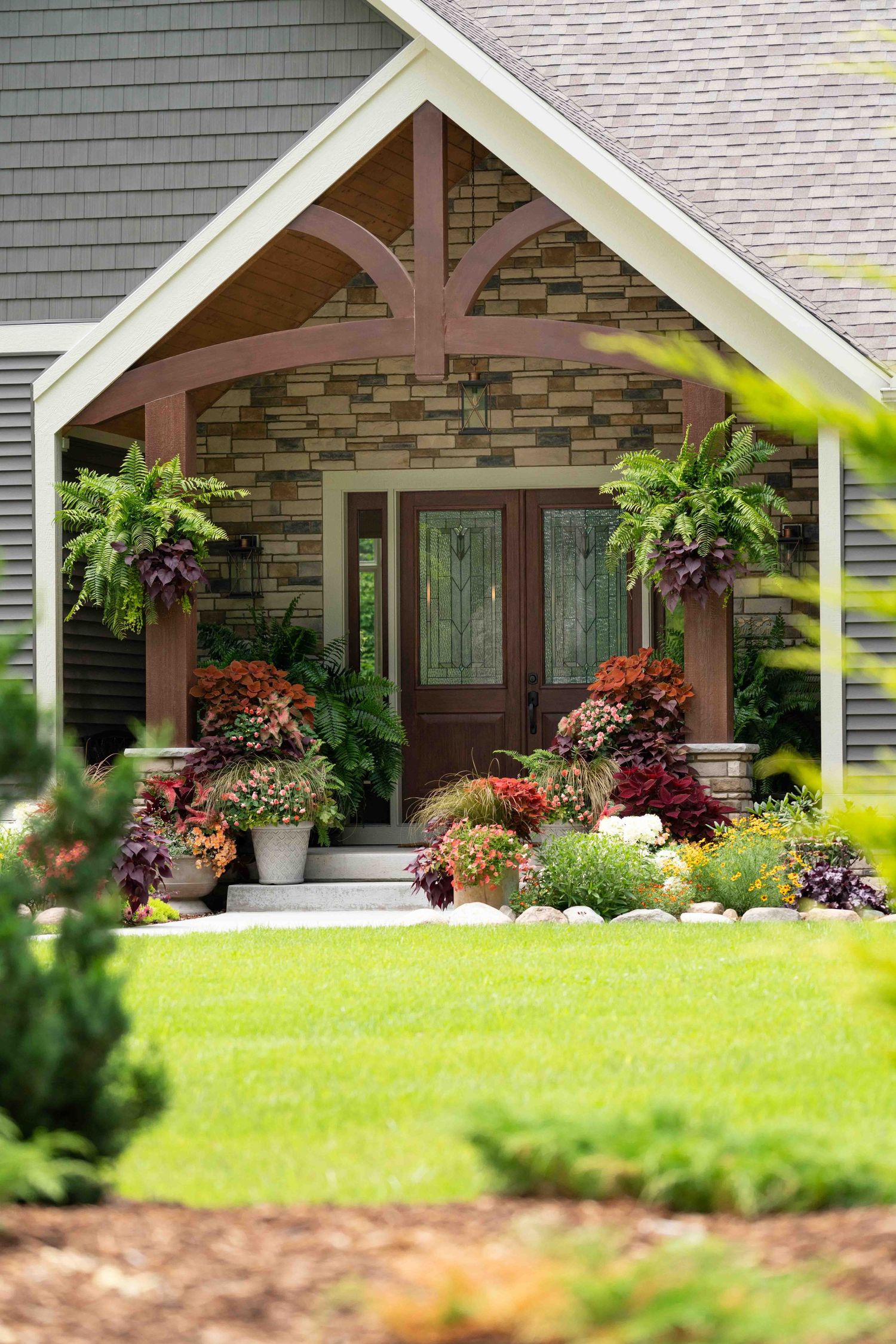 Elegant home entrance with stone facade, potted ferns, and colorful flower arrangements.