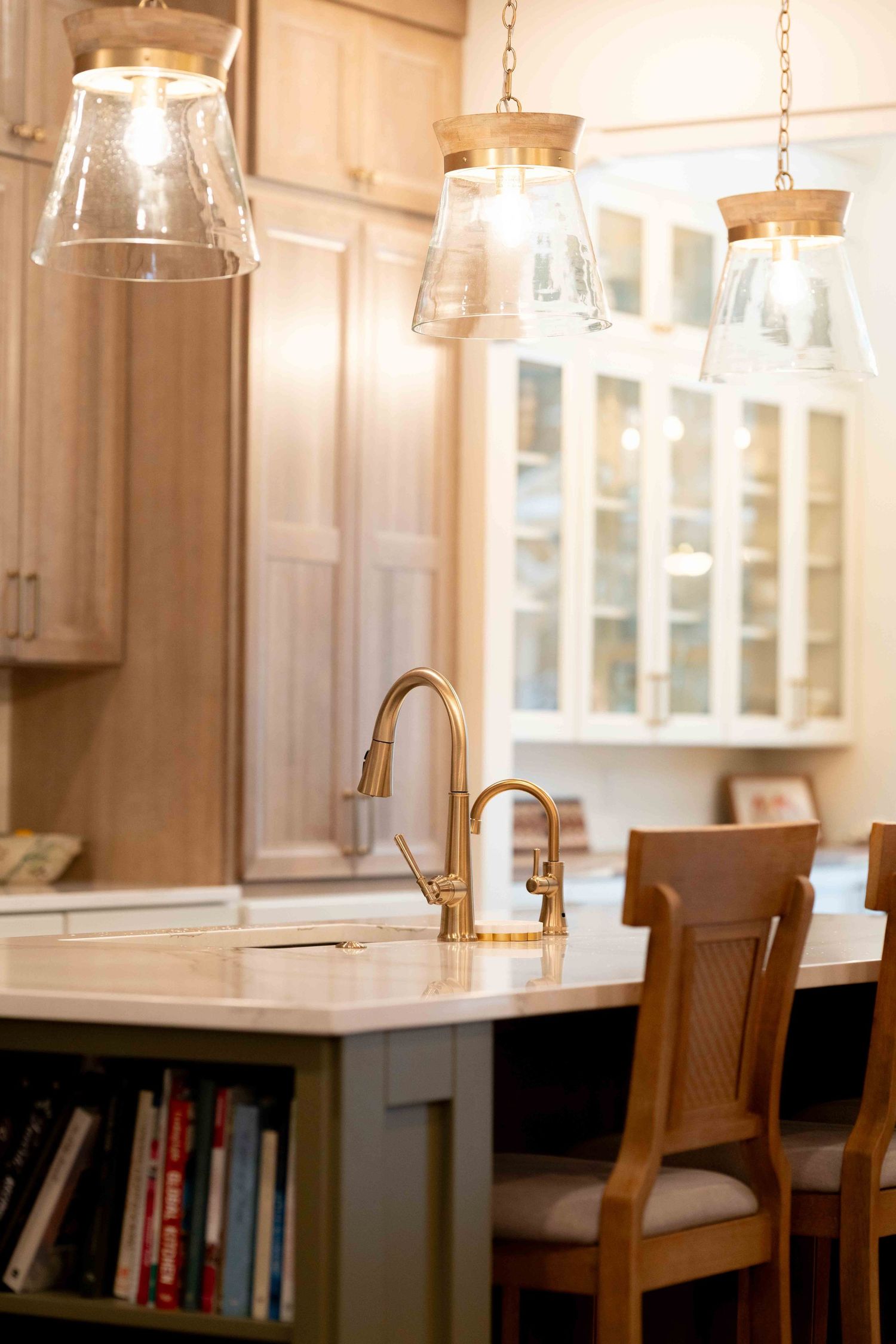 Kitchen island with brass faucet, glass pendant lights, and light wood cabinets with glass doors.