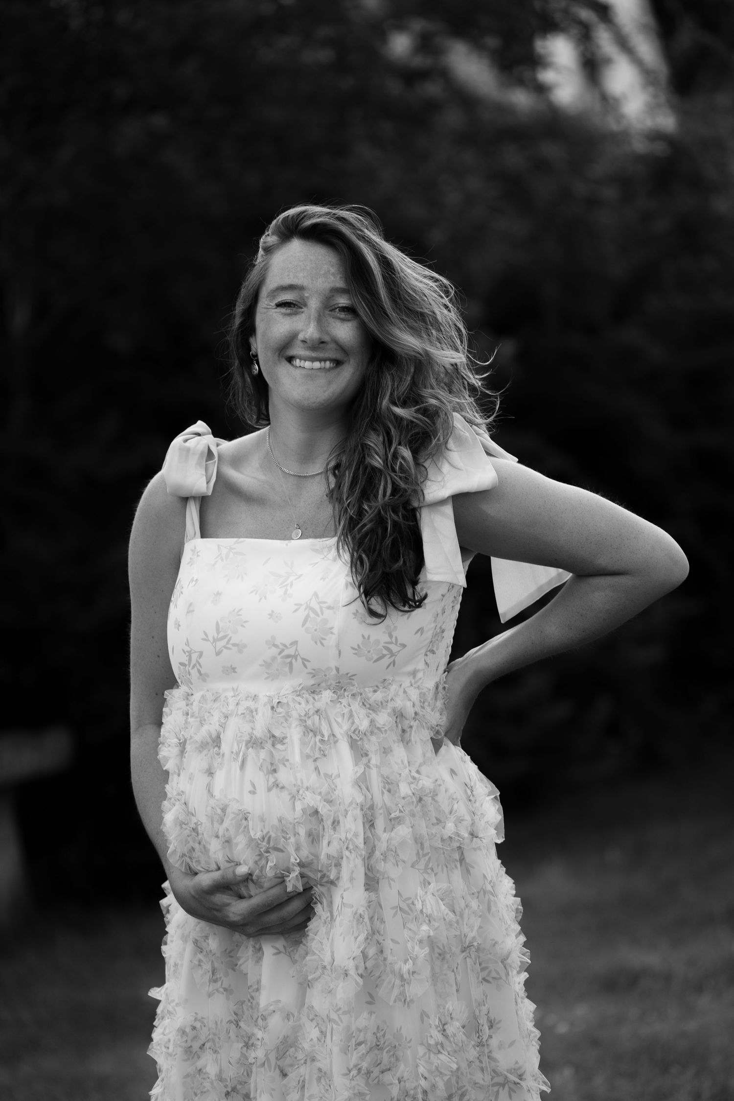 Smiling pregnant woman in white floral dress outdoors in black and white photo.