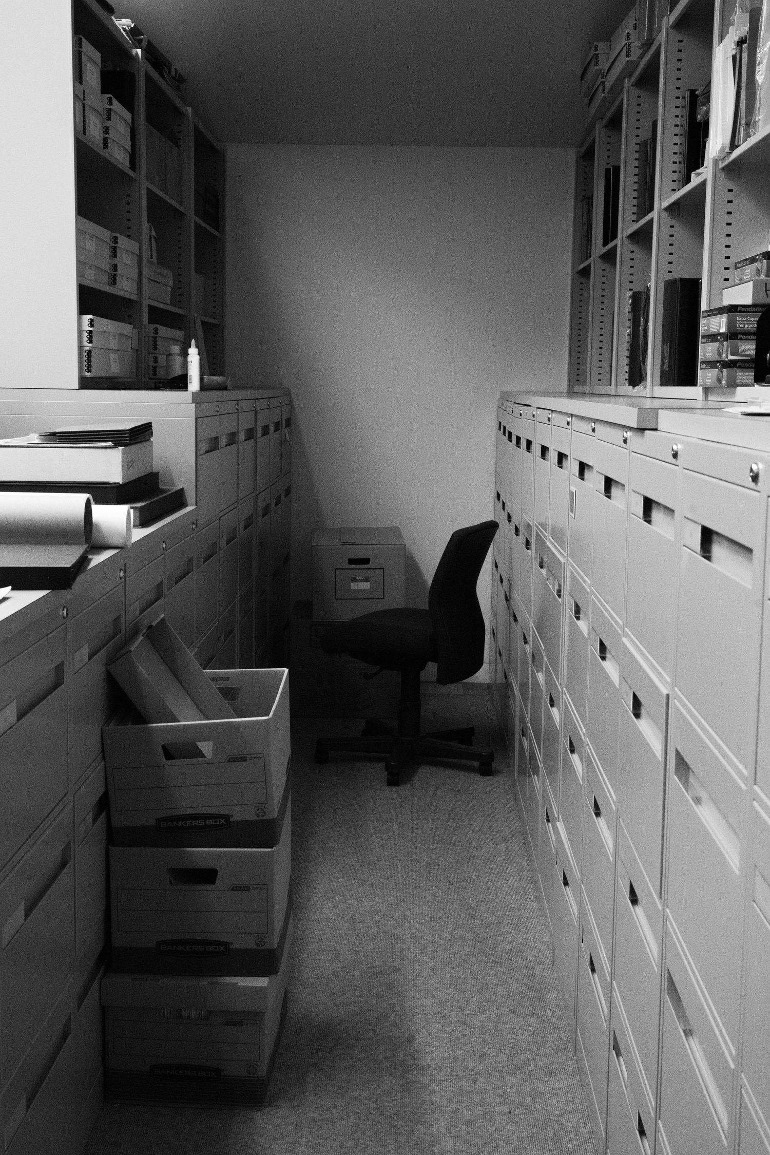Black and white photo of a narrow storage room with shelves, filing drawers, boxes, and an office chair.