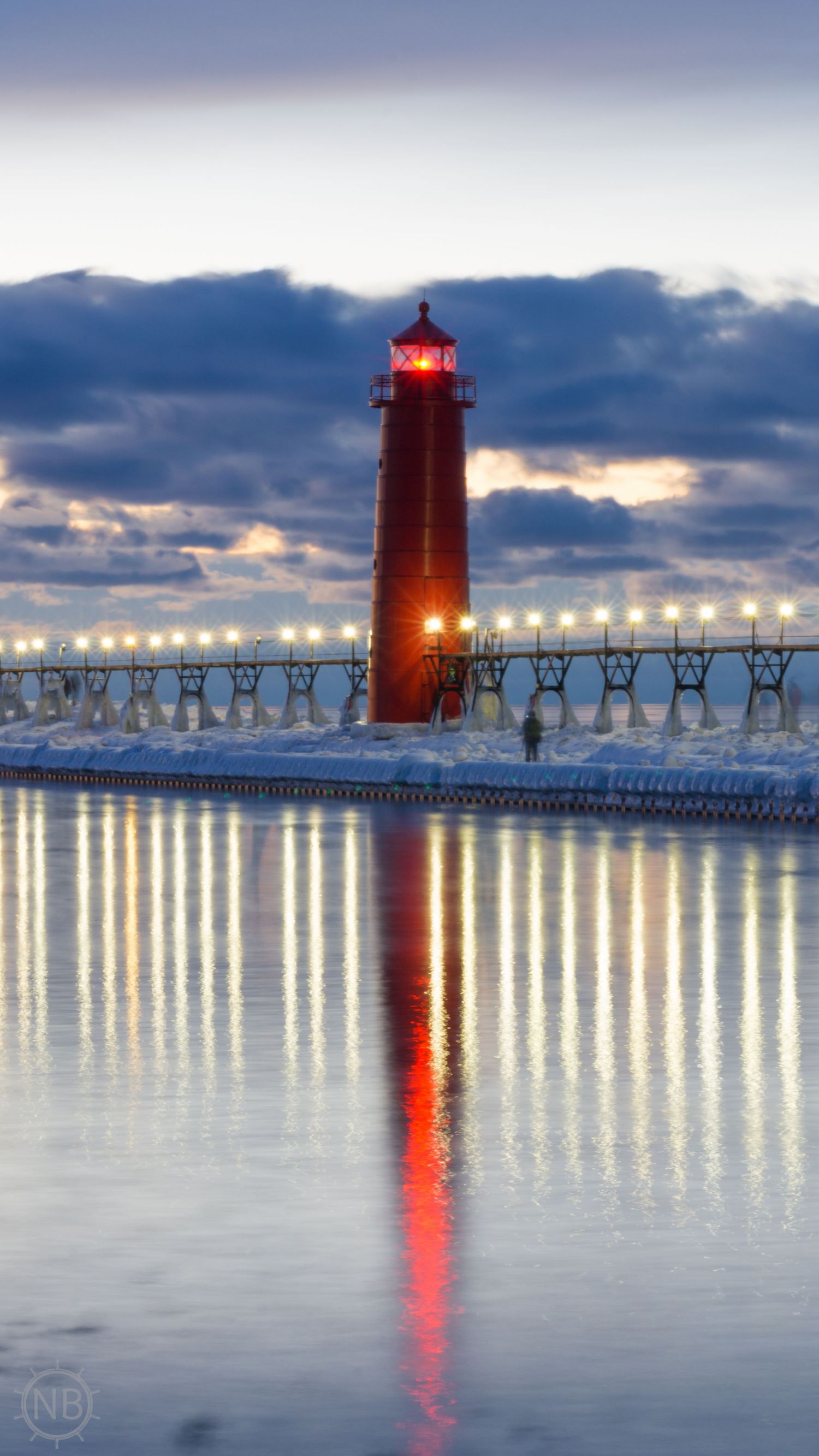 Red Grand Haven lighthouse reflected in calm water at dusk with illuminated pier lights in winter.