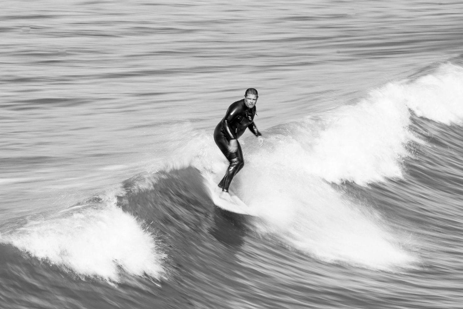 A surfer rides a wave in black and white photography wearing a wetsuit.