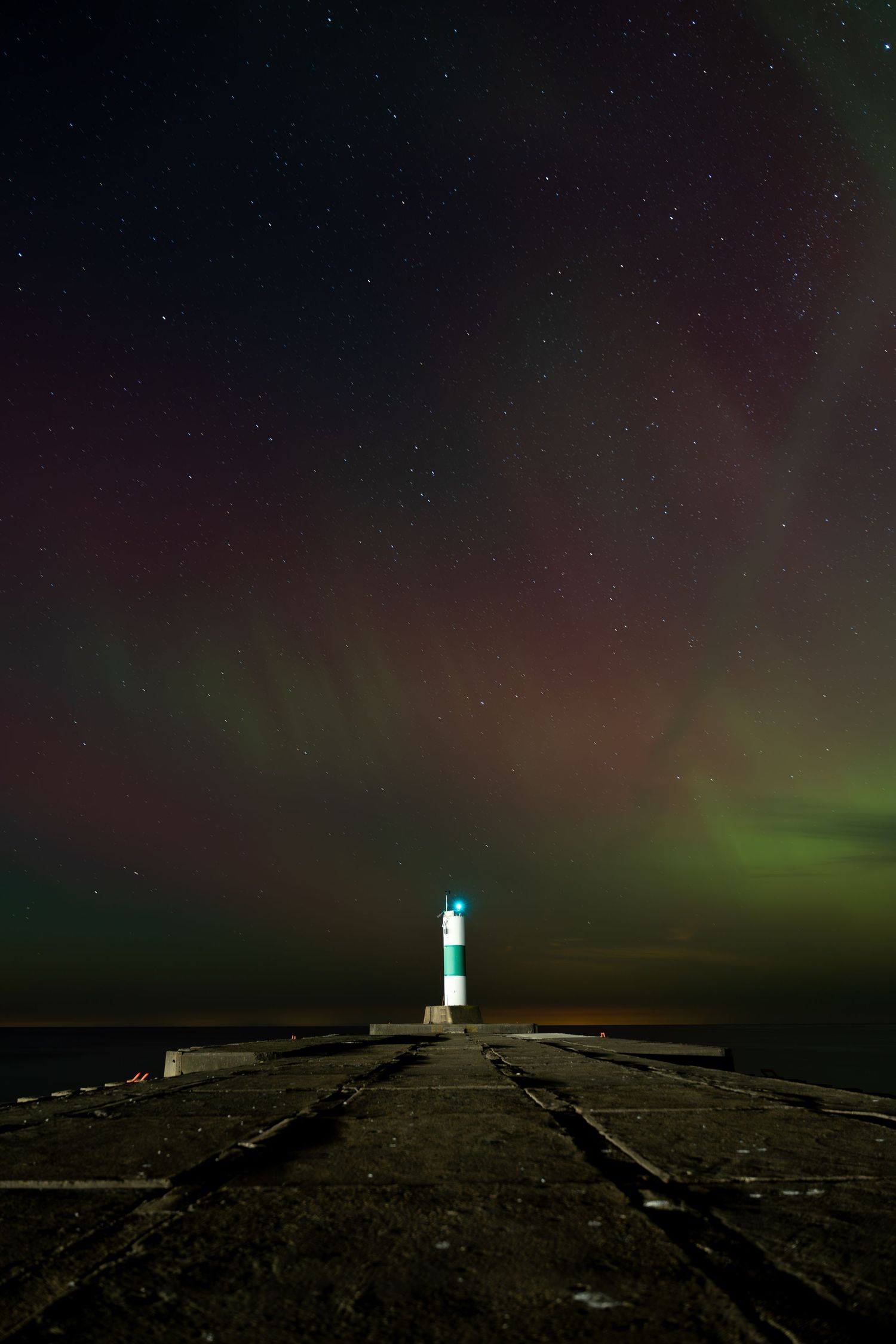 Illuminated lighthouse on a stone pier under a stunning green and red aurora borealis night sky.